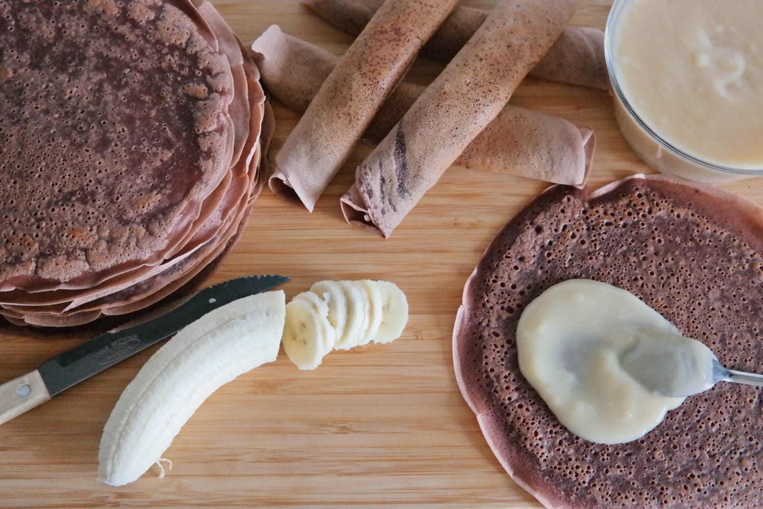 Clătite cu cacao suprapuse și rulate, o clătită cu budincă de vanilie și o banană parțial feliată, văzute de sus