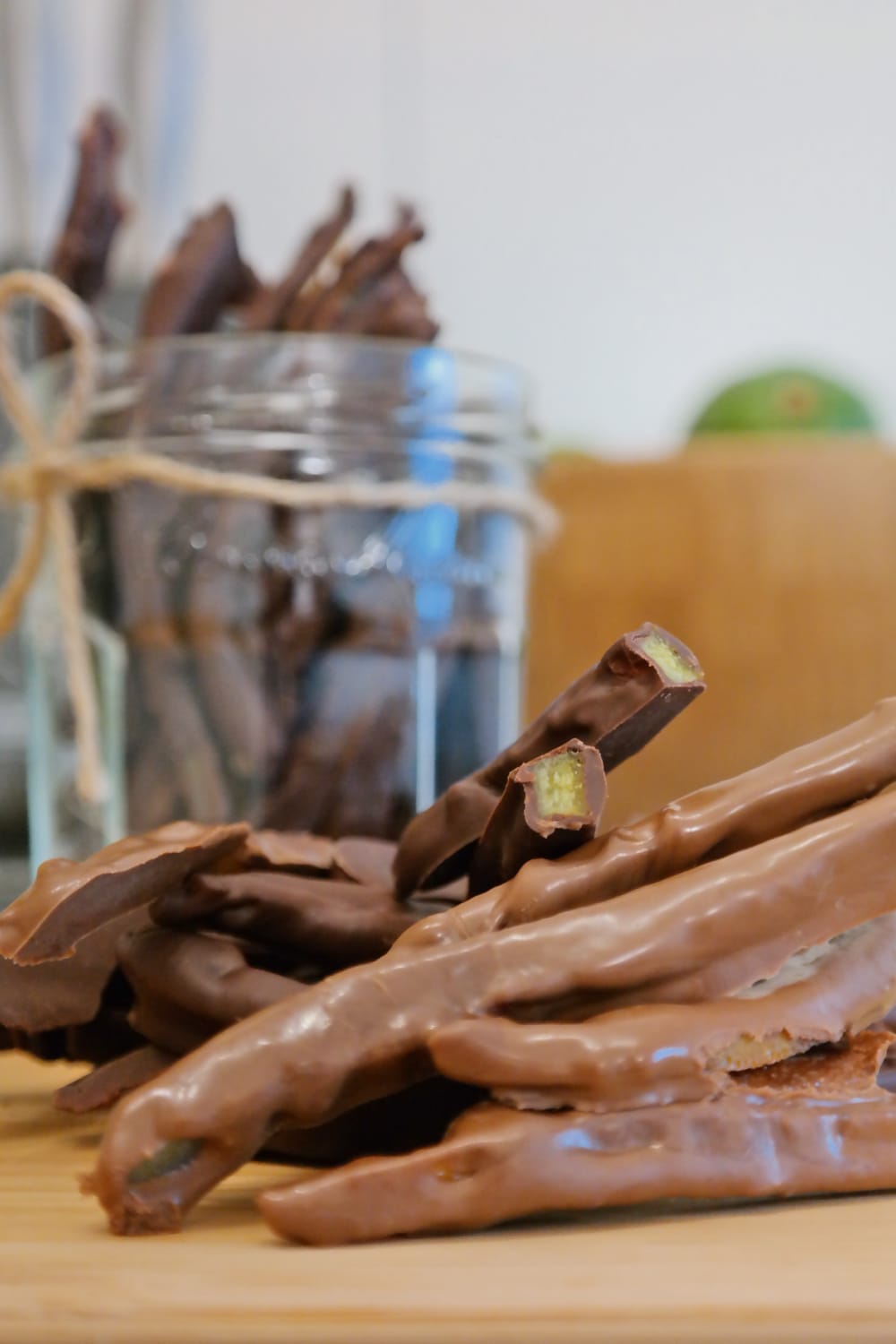Orangettes on a wooden surface, with candied orange peel visible inside two broken pieces and a jar in the background