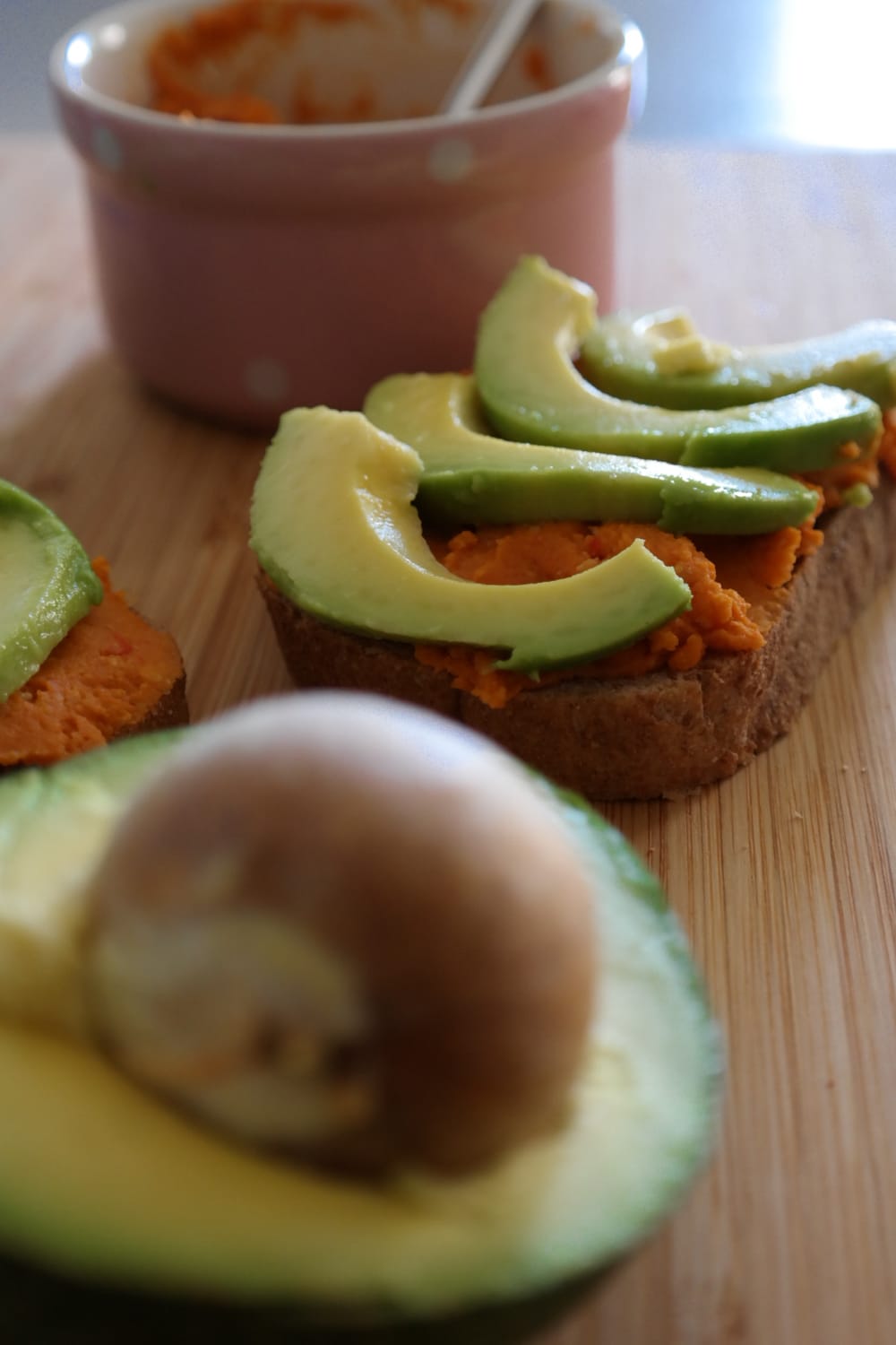 Felii de pâine cu hummus de ardei copt și avocado, cu un bol roz în fundal