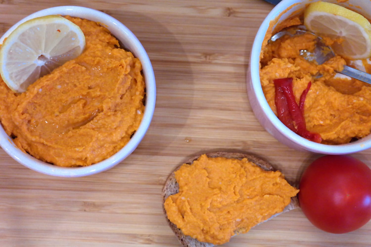 Două boluri roz cu hummus de ardei copt, o felie de pâine unsă cu hummus și o roșie, pe un tocător