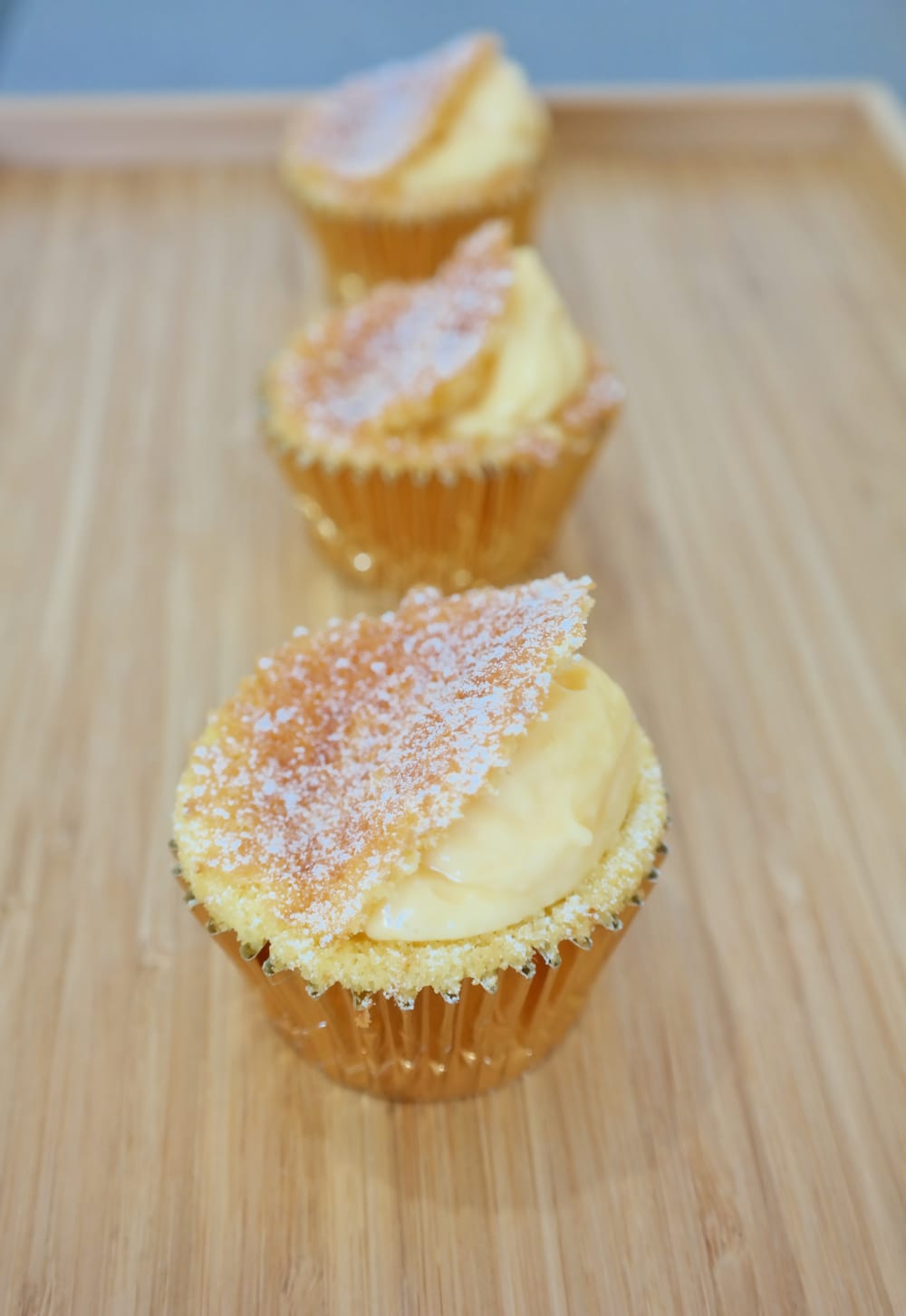 Three orange muffins in gold foil liners, filled with vanilla cream and topped with a sugar-dusted muffin cap, served on a wooden tray