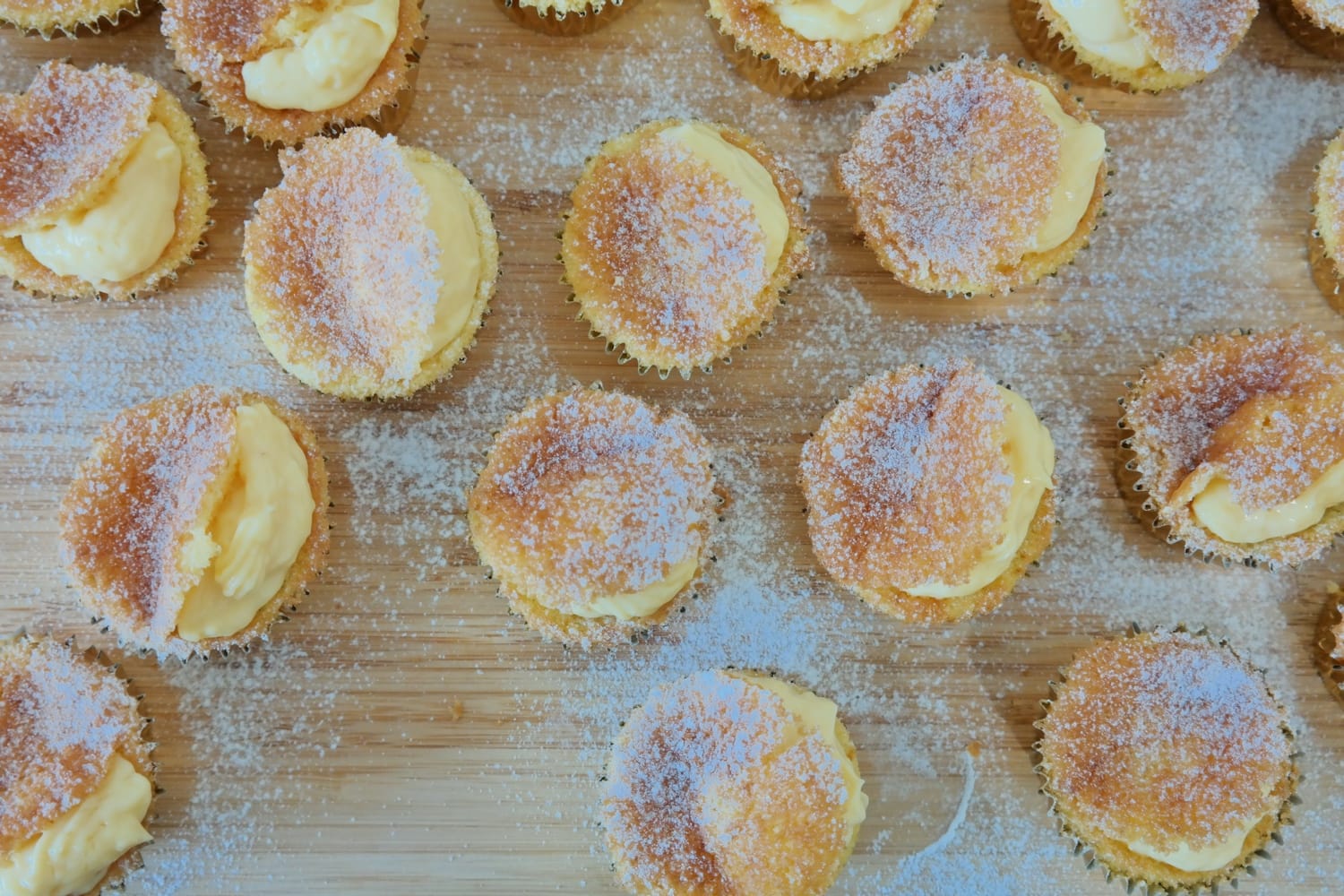 Overhead view of orange muffins filled with vanilla cream, each topped with its partially cut cap and dusted with powdered sugar, on a wooden board