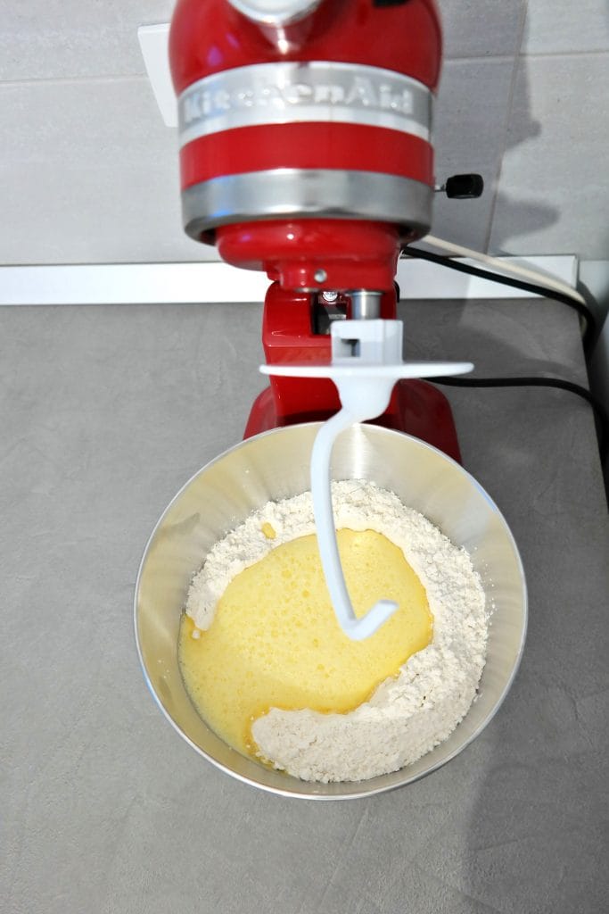 Wet egg and milk mixture poured over flour and dry yeast inside a red KitchenAid stand mixer bowl, ready to knead cinnamon roll dough.