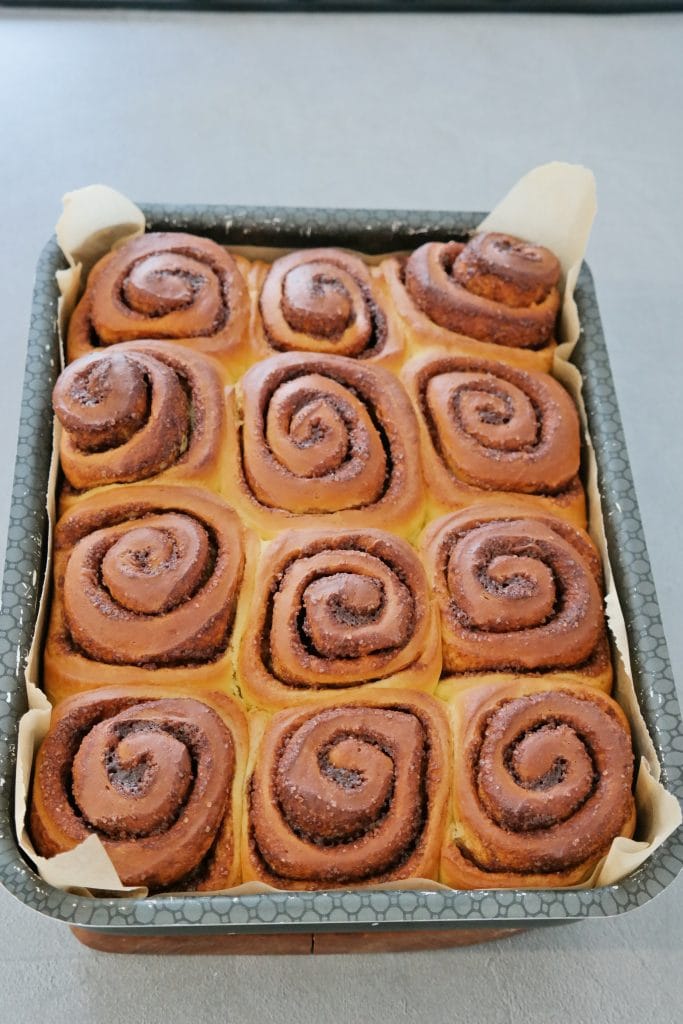 Golden brown freshly baked cinnamon rolls, puffed up and tightly packed inside a parchment-lined rectangular baking pan.