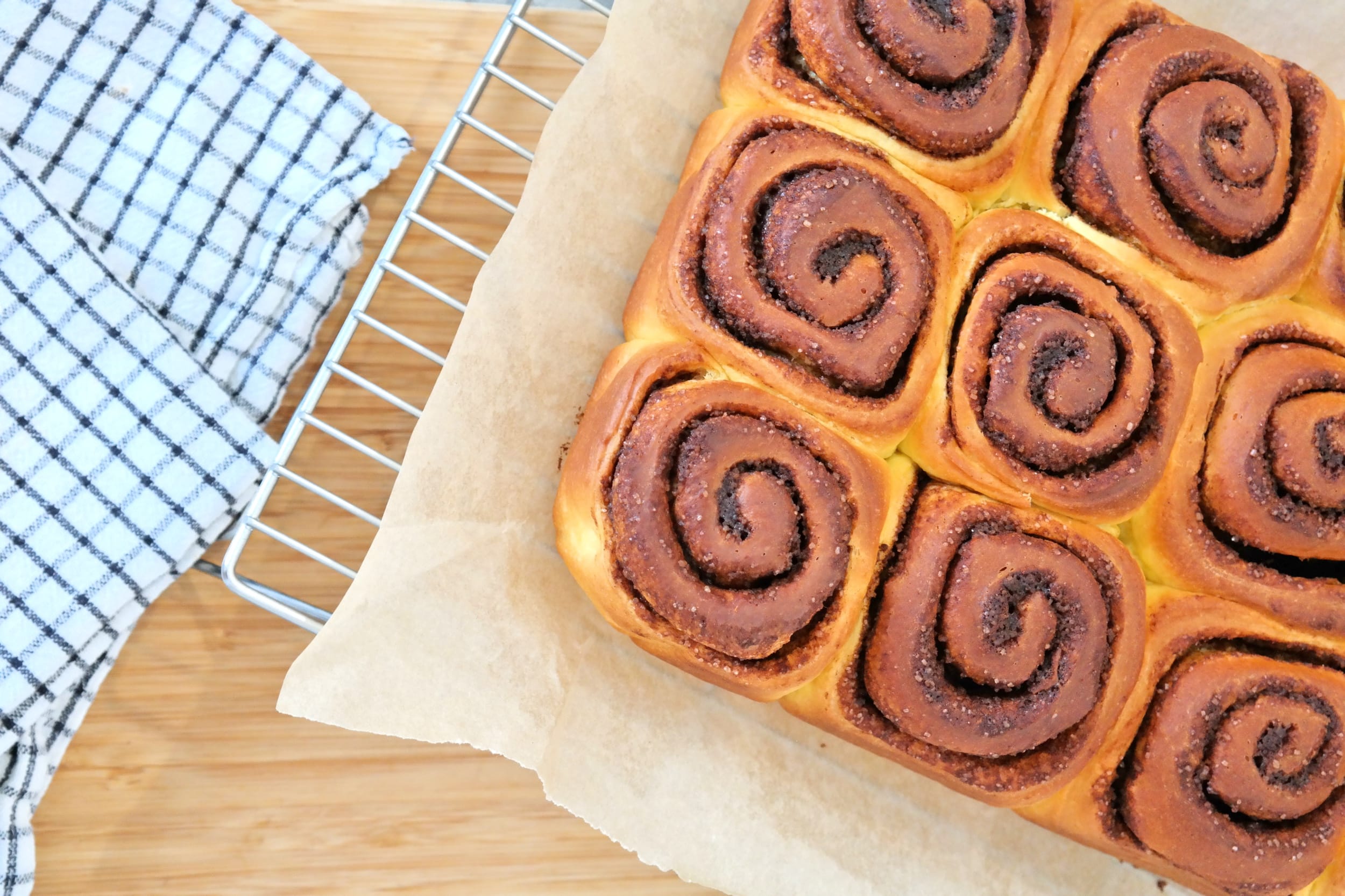 Golden baked cinnamon rolls placed on parchment paper over a metal cooling rack next to a checkered kitchen towel.