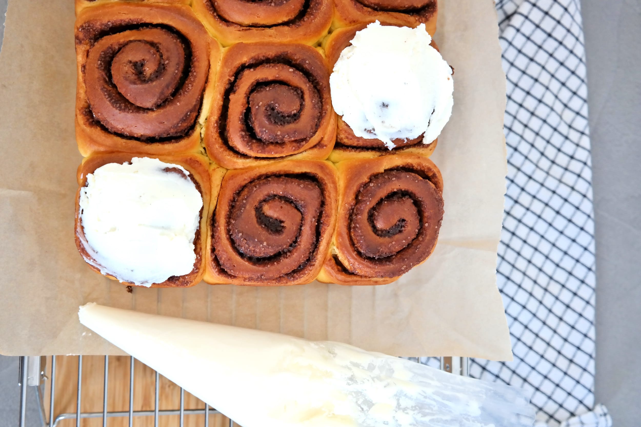 Freshly baked cinnamon rolls on a cooling rack, being decorated with white cream cheese frosting using a pastry piping bag.