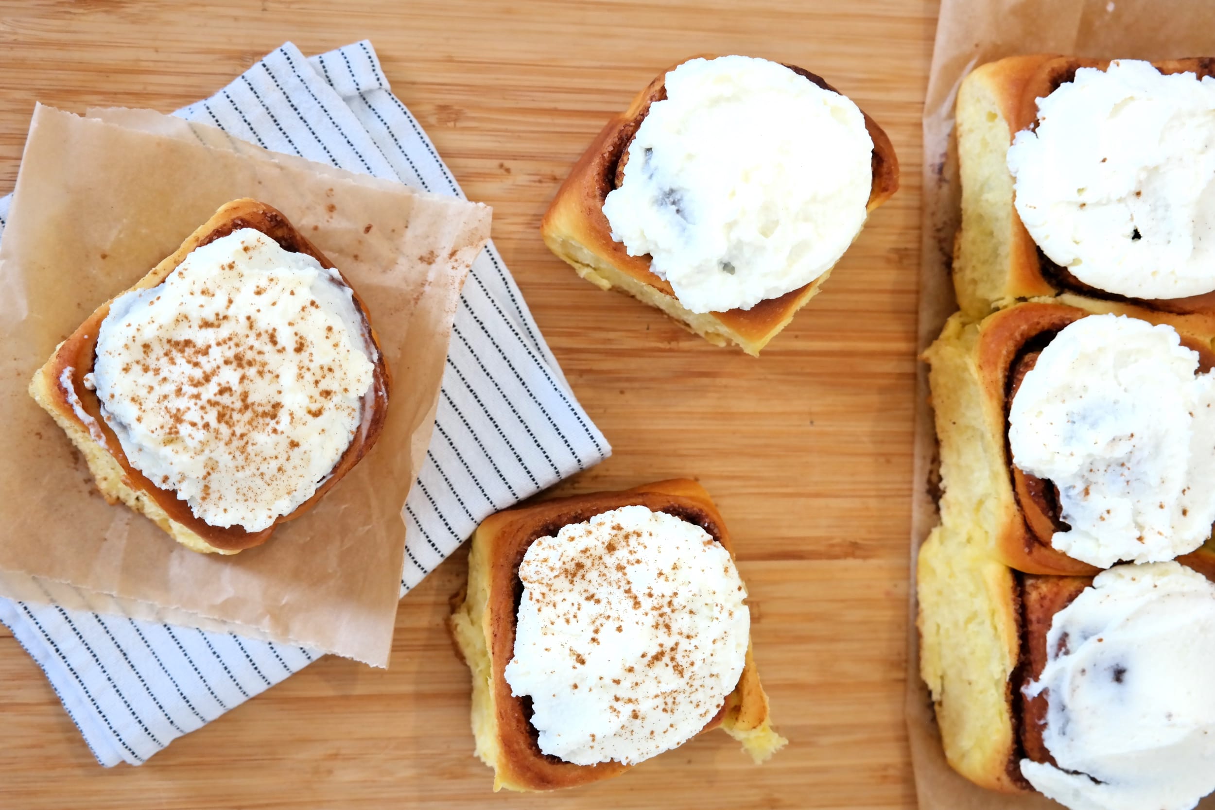 Fresh cinnamon rolls topped with white cream cheese frosting and sprinkled cinnamon, arranged on a wooden cutting board.