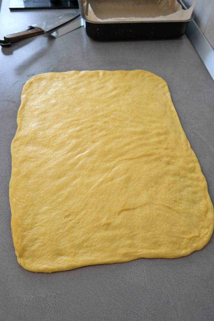 Proofed cinnamon roll dough rolled out evenly with a rolling pin into a large rectangular shape the kitchen countertop, ready for filling.