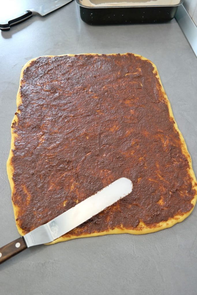 Rectangular cinnamon roll dough evenly covered with butter, sugar, and cinnamon paste using an offset metal spatula on a countertop.