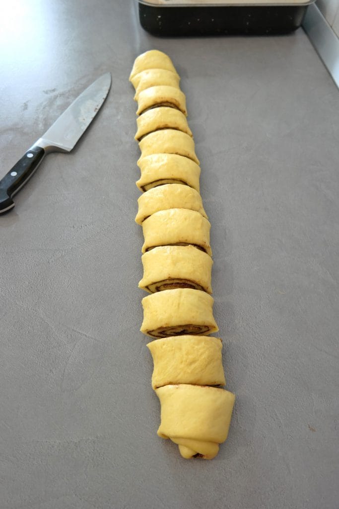 Cinnamon roll dough log sliced into individual portions on a grey countertop, placed next to a sharp chef knife.