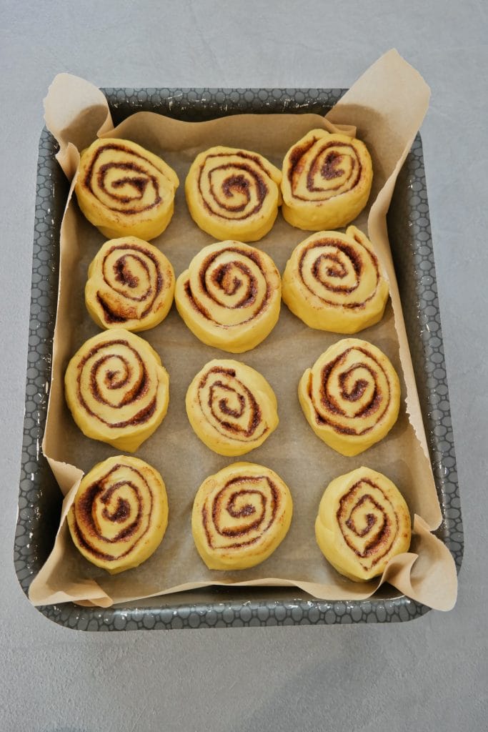 12 unbaked cinnamon rolls arranged on parchment paper in a baking pan, ready for their second proofing before baking.