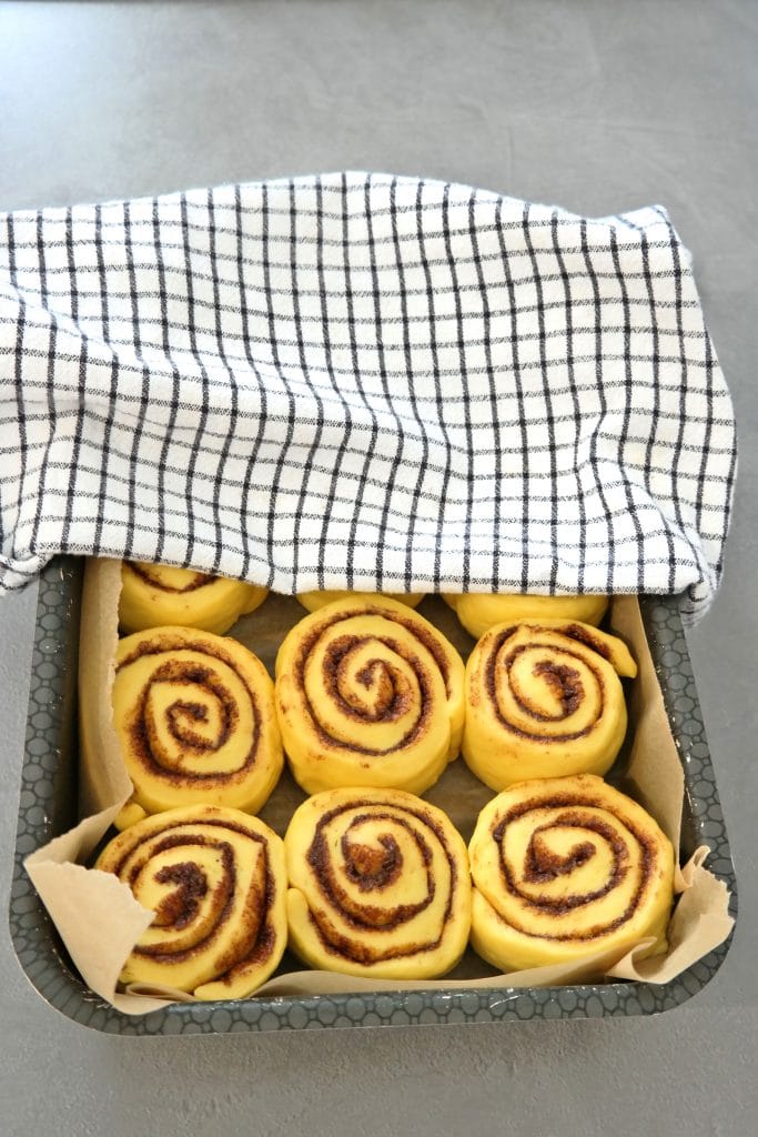 Baking pan with cinnamon rolls partially covered with a kitchen towel, resting for their second proofing before baking.