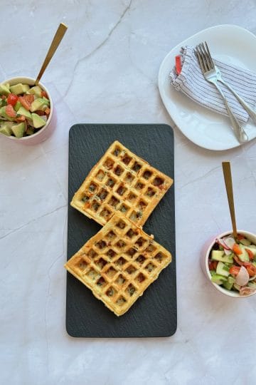 Vedere de sus cu două gofre sărate pe un platou negru, între două boluri roz cu salată de avocado și legume