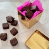 Gold gift box lined with bright pink tissue paper and filled with chocolate-covered brownie bites, with several more brownie bites topped with sea salt scattered on a marble surface nearby.