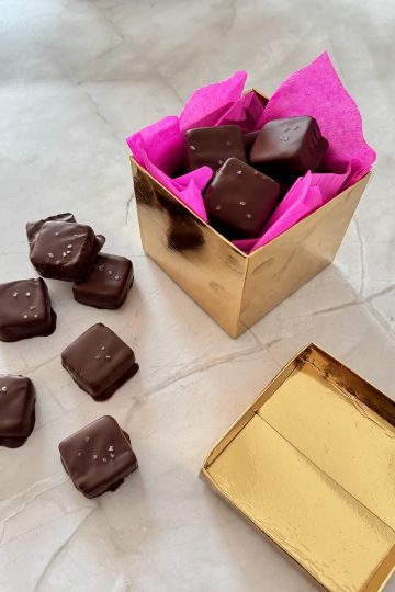 Gold gift box lined with bright pink tissue paper and filled with chocolate-covered brownie bites, with several more brownie bites topped with sea salt scattered on a marble surface nearby.