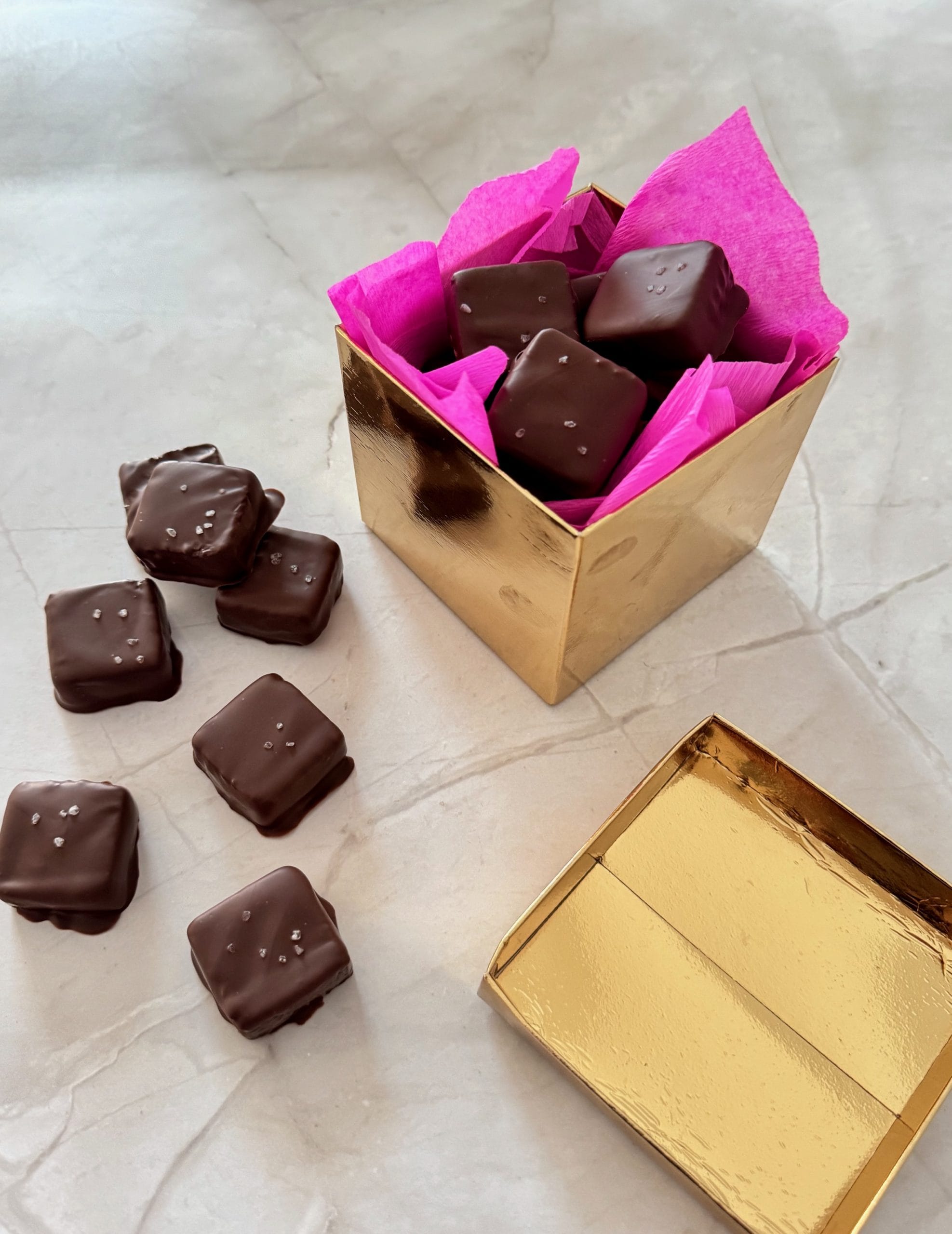 Gold gift box lined with bright pink tissue paper and filled with chocolate-covered brownie bites, with several more brownie bites topped with sea salt scattered on a marble surface nearby.