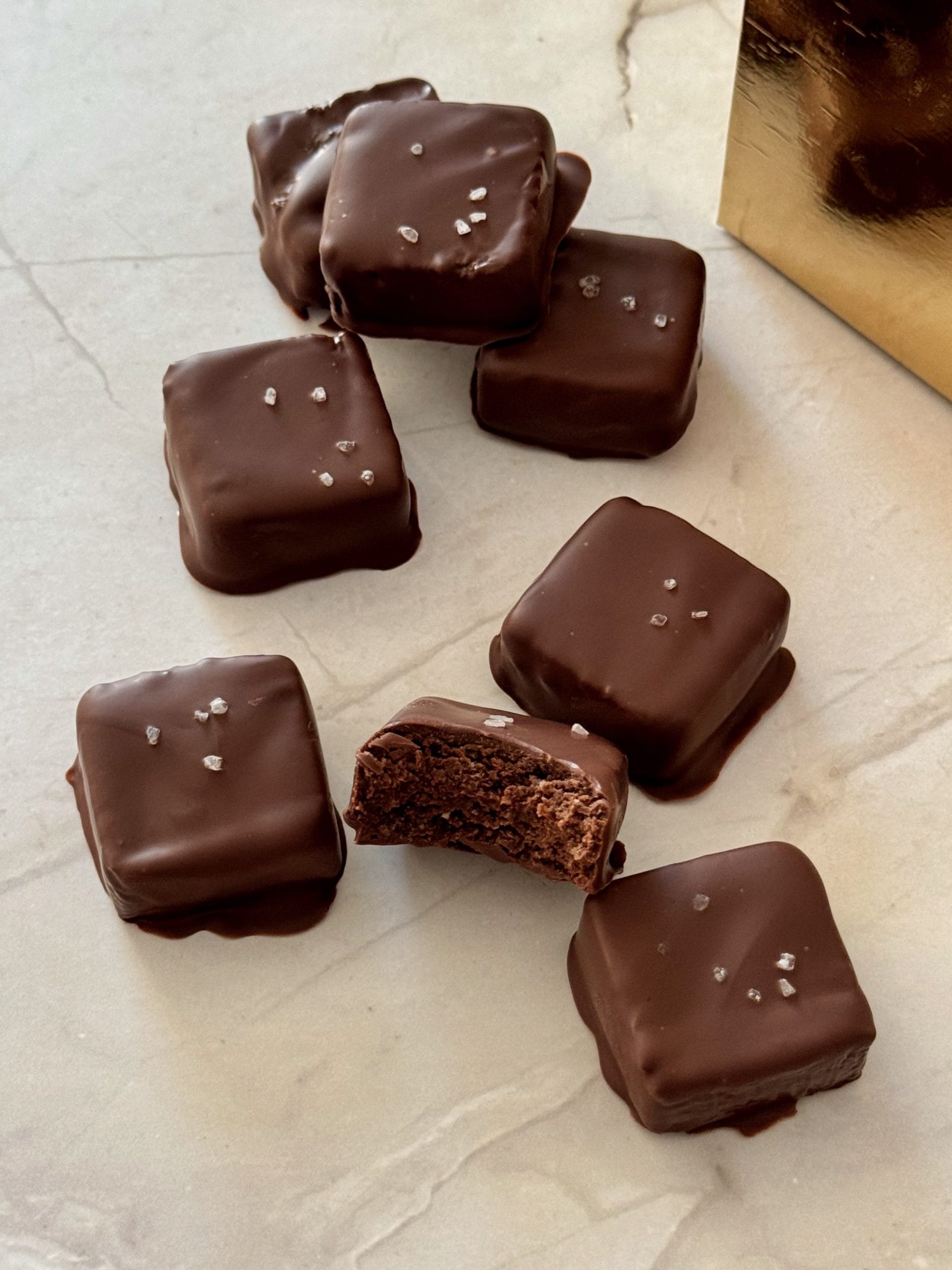 Bite-sized chocolate-covered brownies topped with sea salt, with one brownie showing a rich, fudgy center, arranged on a marble surface beside a gold box.