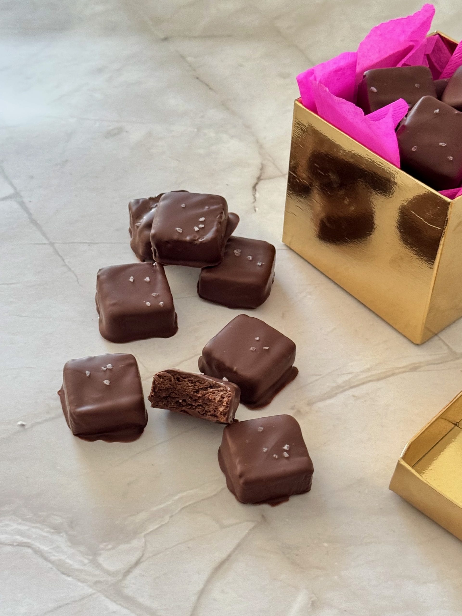 Small square chocolate-covered brownie bites sprinkled with sea salt, displayed on a marble surface next to a gold gift box lined with pink tissue paper.