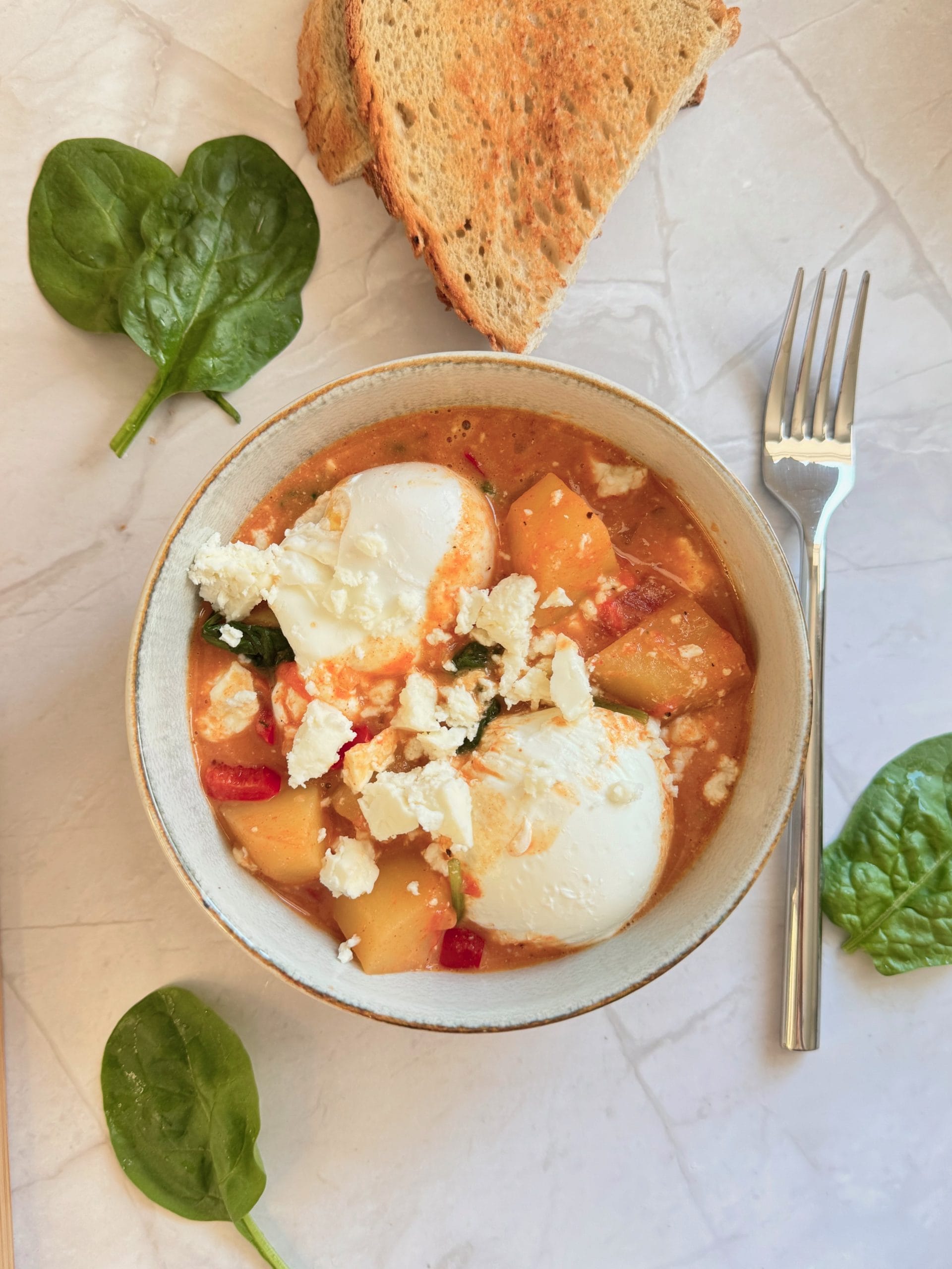 A serving bowl of potato and spinach shakshuka with poached eggs and feta