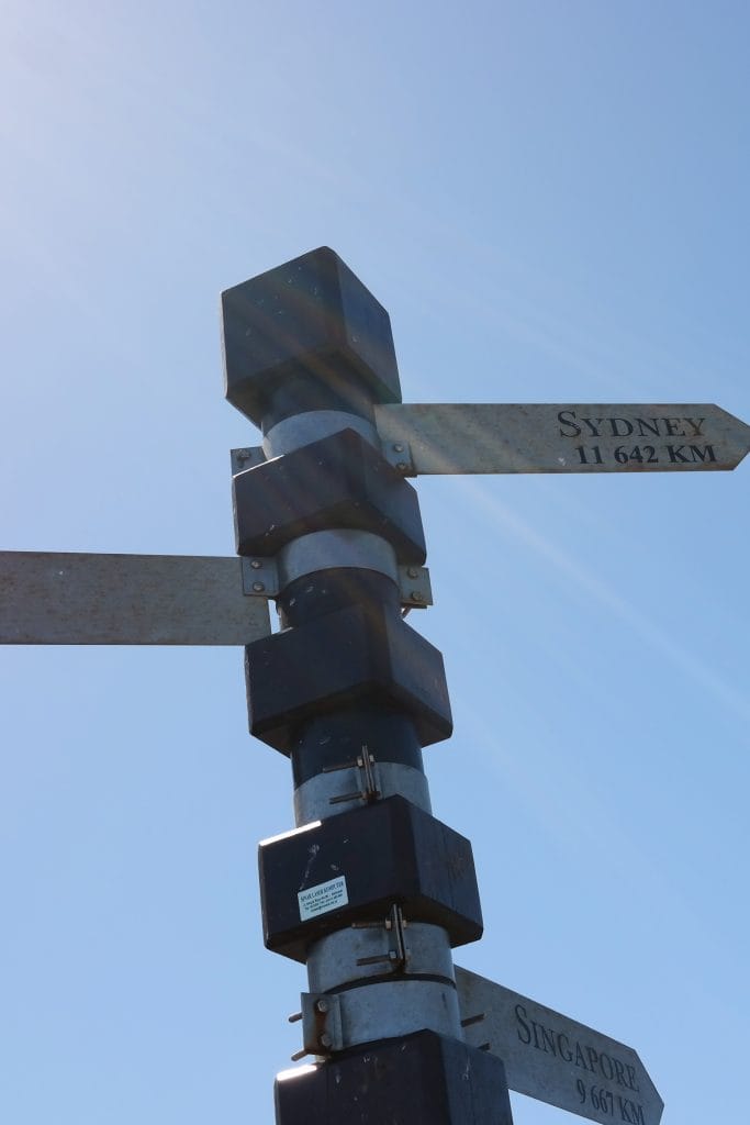 O sectiune a stalpului indicator cu sageti, pe care se distinge sageata spre Sydney cu distanta de 11.642 km, sub cerul senin cu razele soarelui in Cape Town.