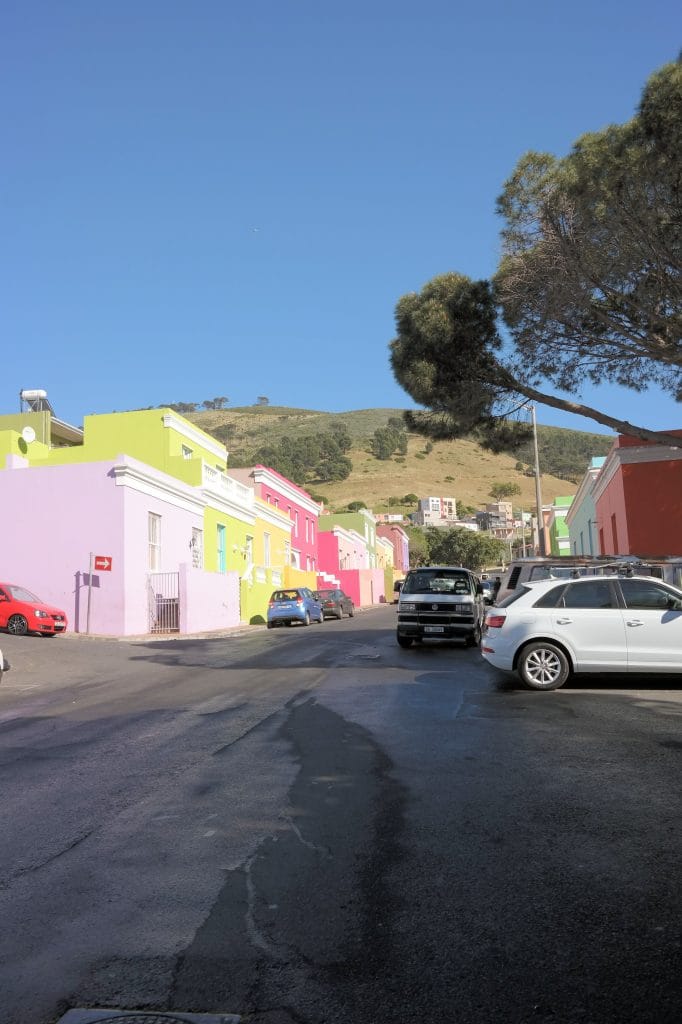 O strada asfaltata dintr-un cartier cu case colorate in nuante pastel de roz, galben si verde, cu masini parcate si un deal verde in fundal, in zona Bo-Kaap din Cape Town.