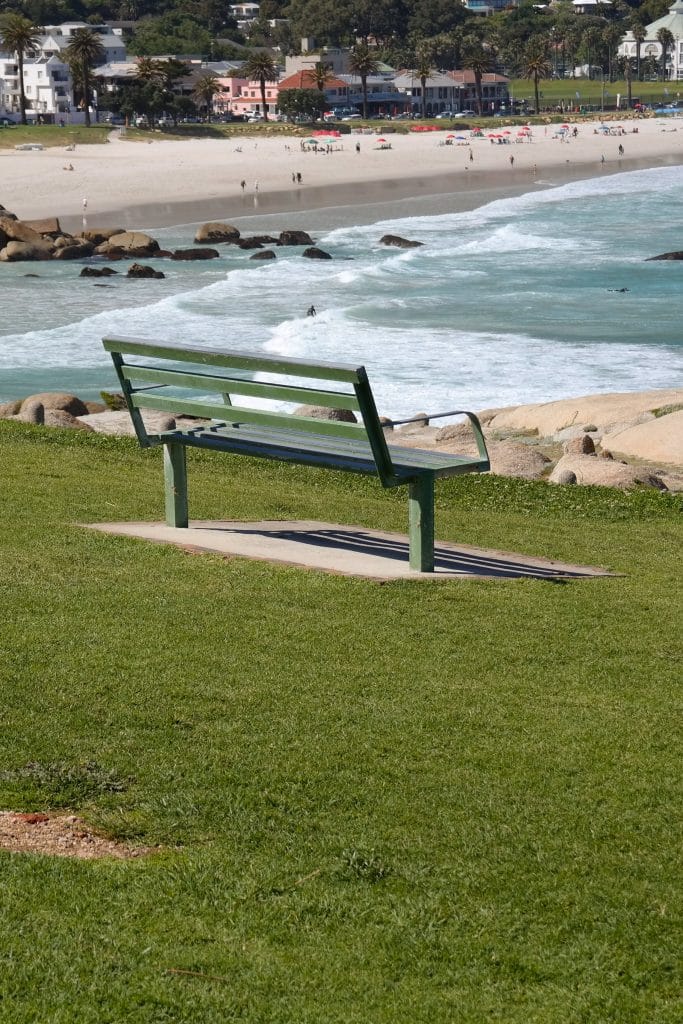 O banca verde amplasata pe o iarba verde pe o colina, orientata spre o plaja cu nisip alb, stanci si apele albastre ale oceanului in Cape Town.