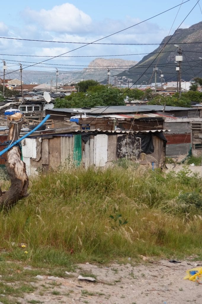 Vedere asupra unei asezari informale dintr-un township in Cape Town, cu locuinte modeste din lemn si tabla, retele de fire electrice si munti in fundal.