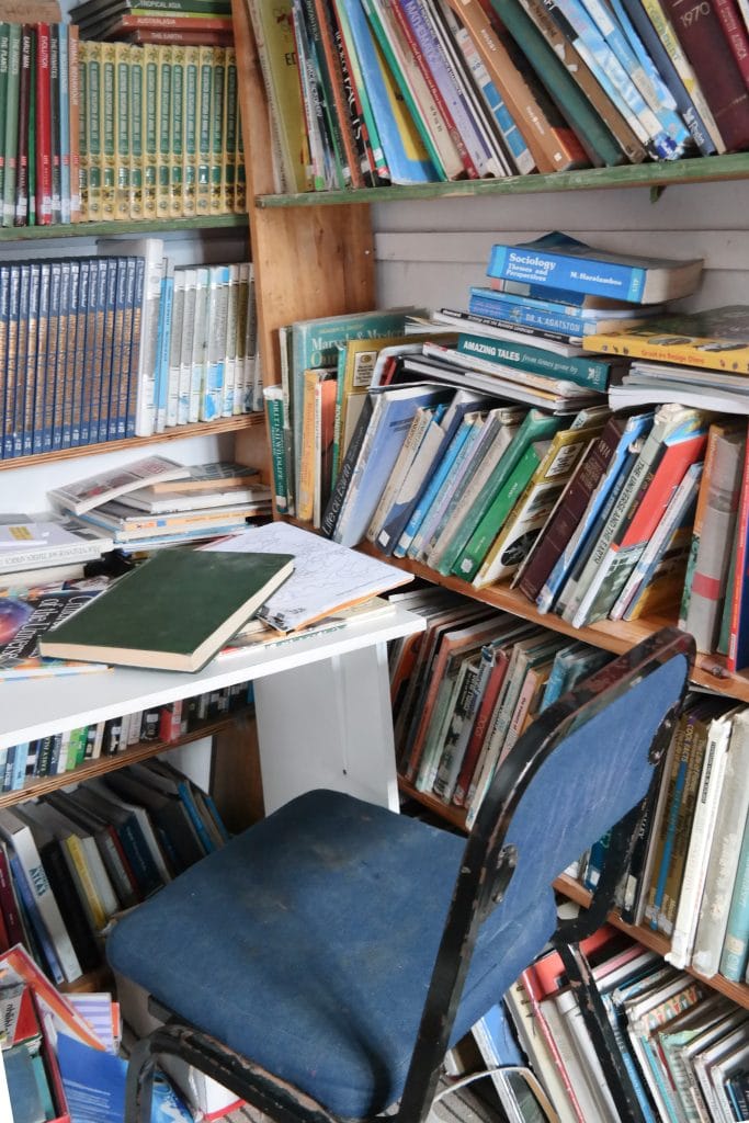 Rafturi de lemn pline cu carti si manuale intr-o biblioteca sau sala de studiu din township, cu un scaun albastru in prim-plan, surprinse in timpul unei vizite in Cape Town.