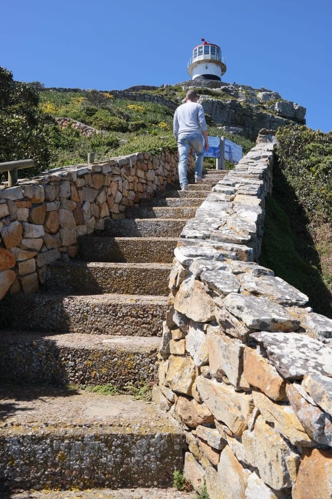Trepte din piatra cu ziduri laterale rustice care duc in sus spre un far alb cu acoperis rosu pe o colina in Cape Town.