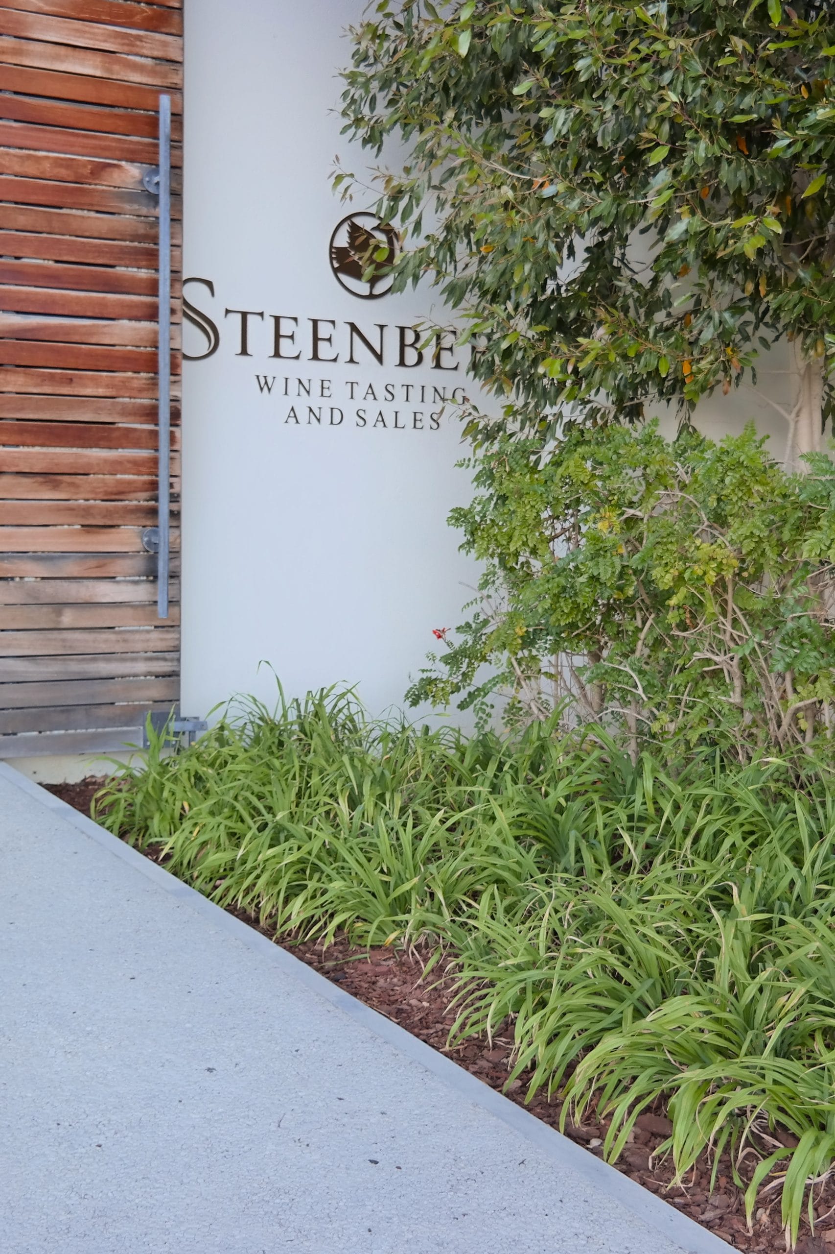 Intrarea moderna in cladirea Steenberg Wine Tasting and Sales, flancata de panouri din lemn si vegetatie verde bogata.