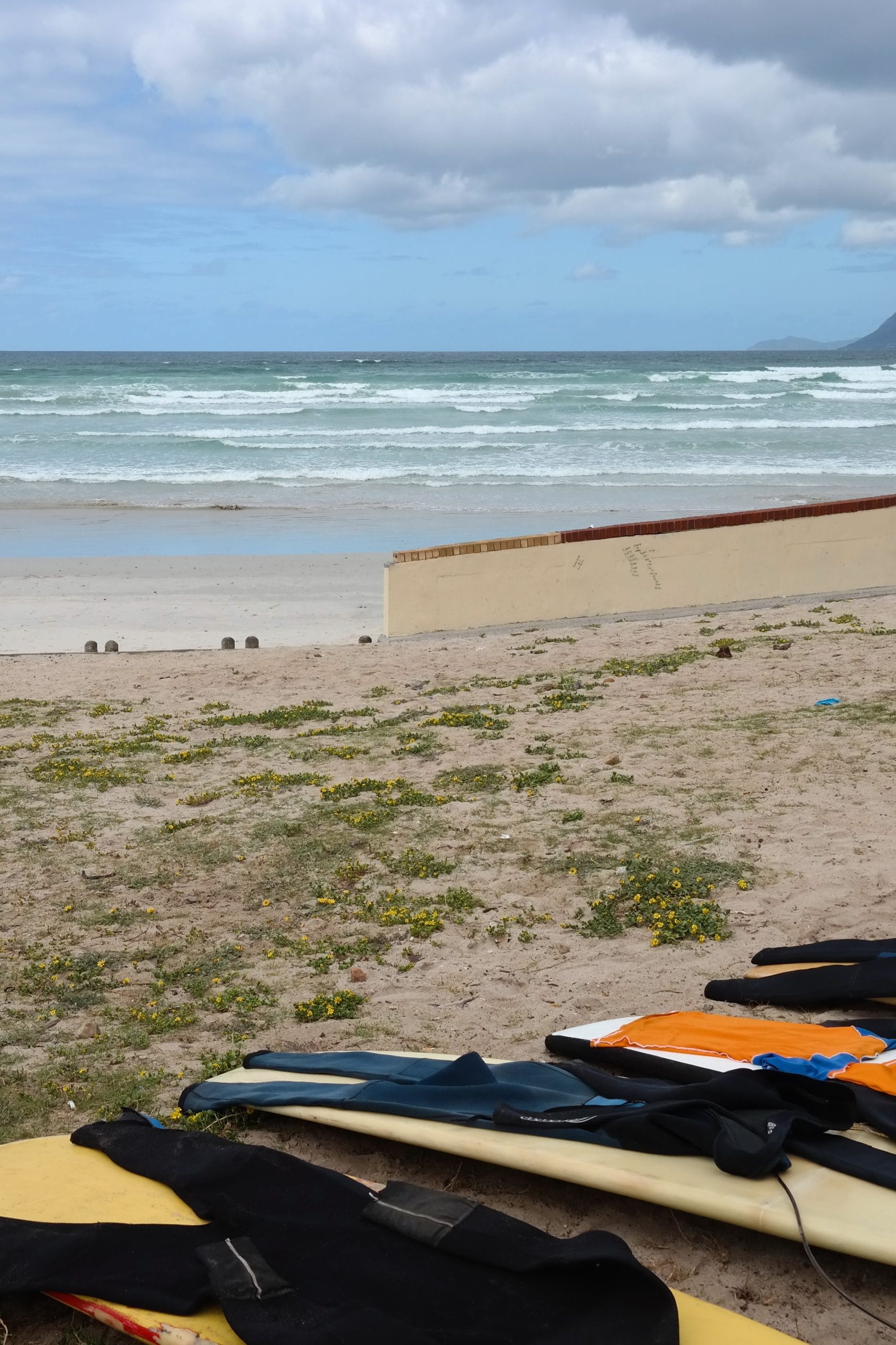 Placi de surf si costume de neopren pe nisip, pregatite in cadrul unui proiect de voluntariat de surf pe o plaja din Cape Town, cu oceanul in fundal.