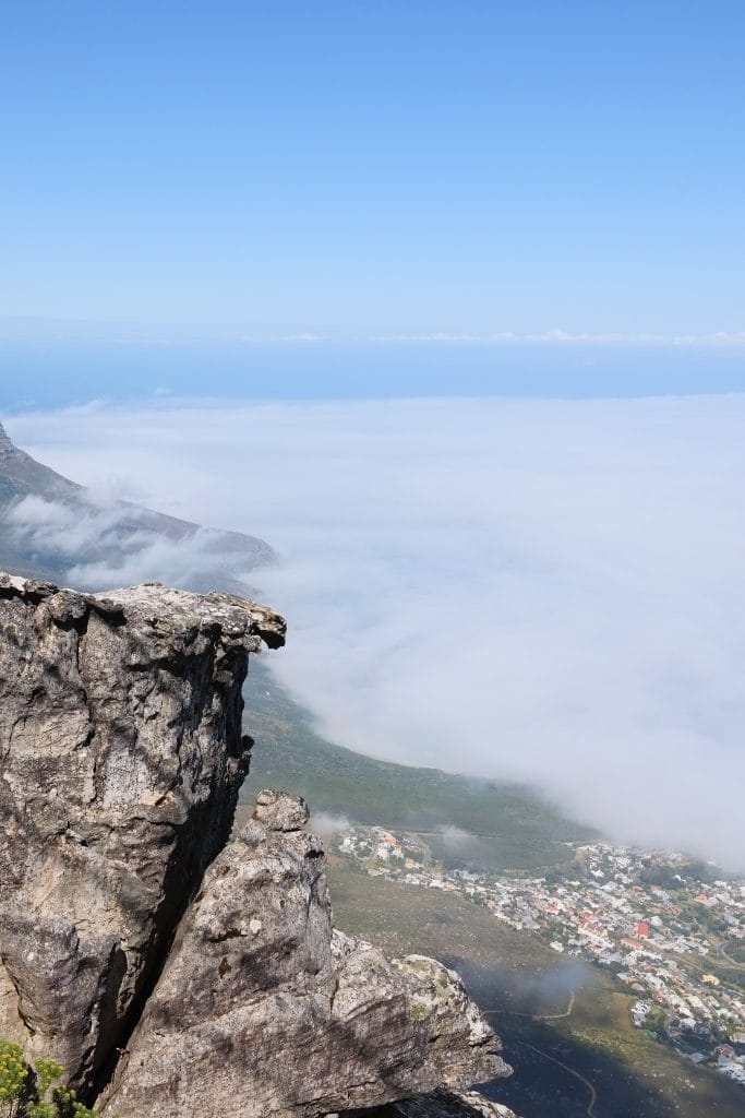 O vedere de pe varful Table Mountain, prezentand stanci masive in prim-plan si un strat dens de nori care acopera orasul si coasta in departare.