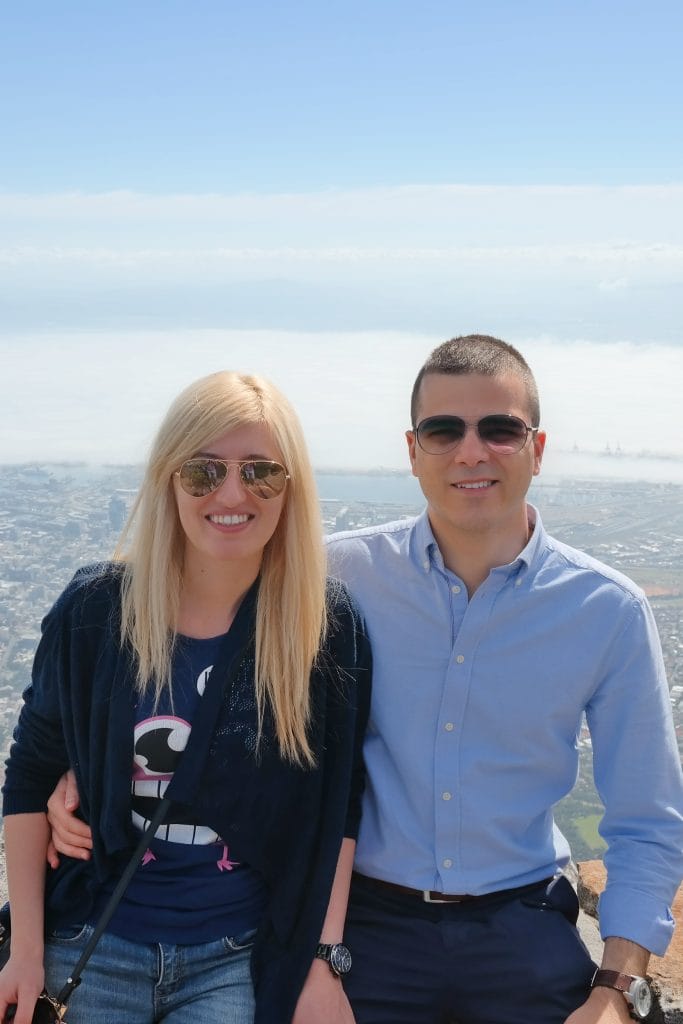 O fotografie de cuplu pe varful Table Mountain, cu un peisaj urban intins si un strat de nori dens in fundal sub un cer senin.