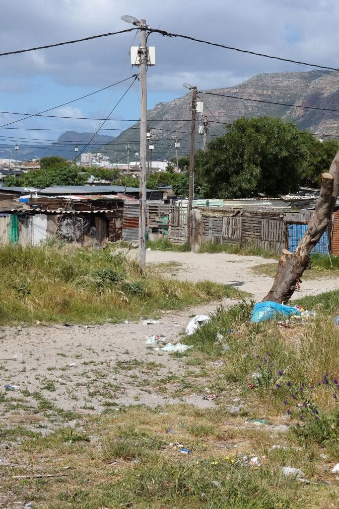 Asezare informala de tip township in Cape Town, cu case modeste construite din tabla si lemn, stalpi de electricitate si un munte impunator in fundal, vazute de pe o carare de pamant.