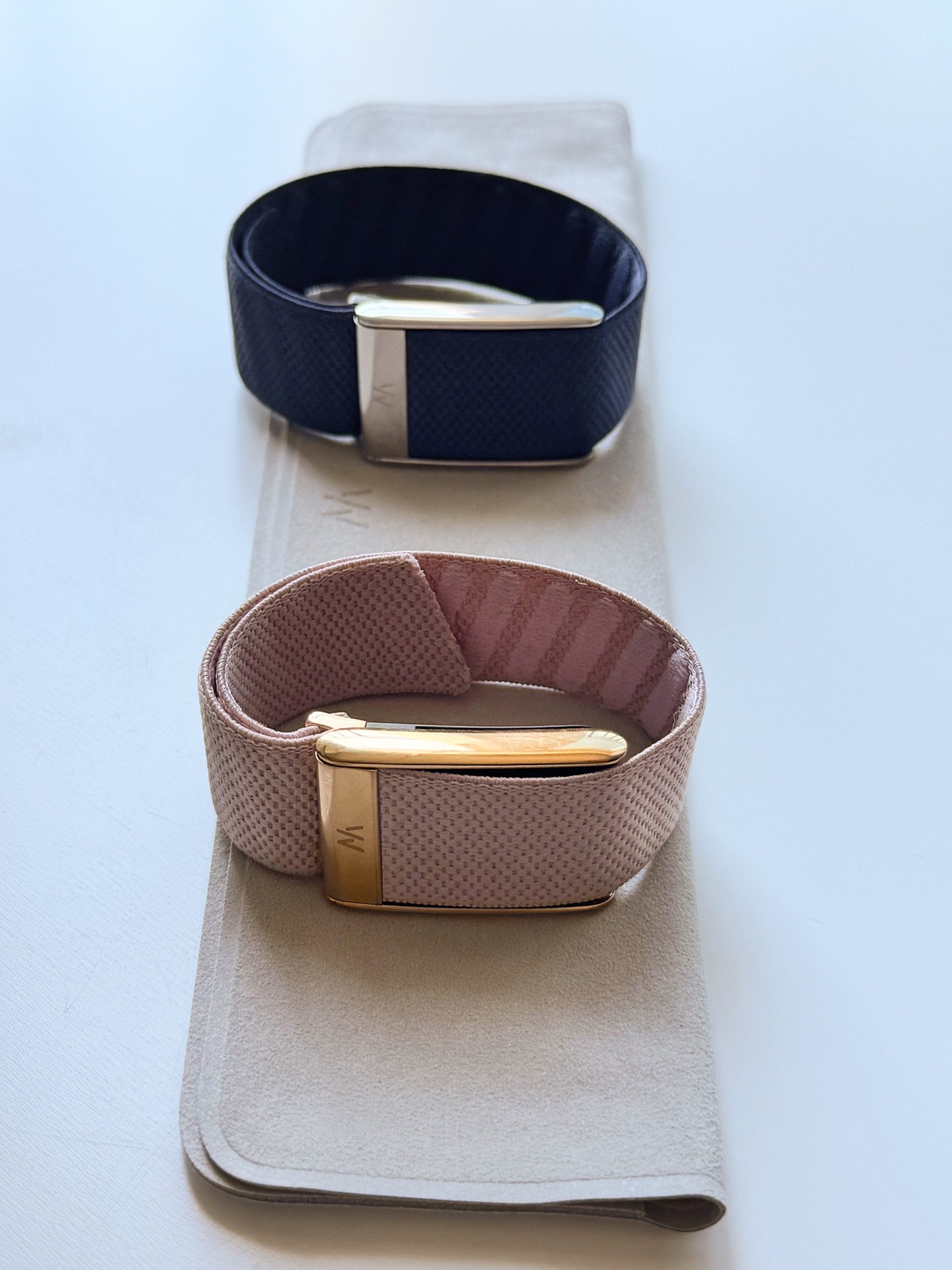 Doua bratari Whoop cu accesorii textile bleumarin si roz, asezate pe un suport deschis la culoare