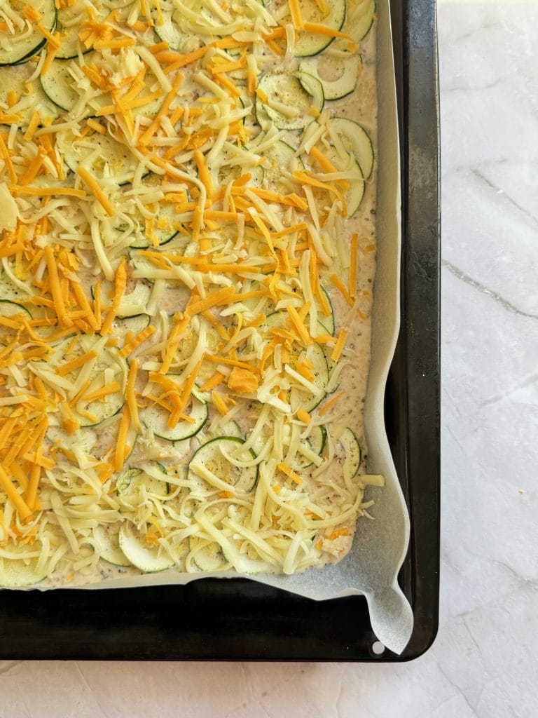 Clatita cu zucchini necoapta, acoperita cu felii subtiri de zucchini, cascaval si cheddar ras, pe o tava de copt