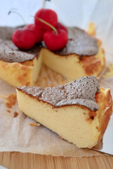 Close-up of a creamy Basque burnt cheesecake slice with a caramelized dark crust and fresh red cherries in the background