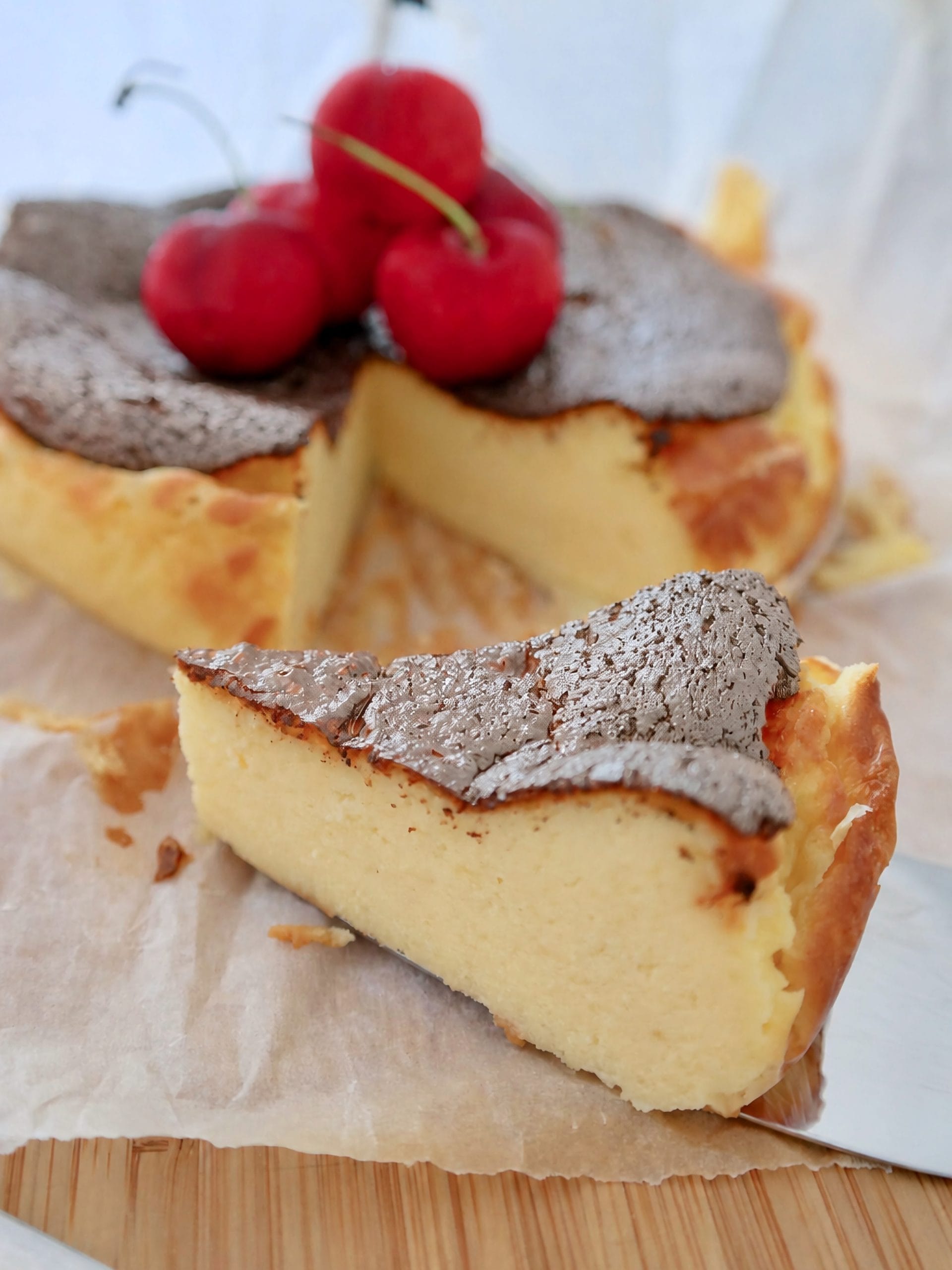 Close-up of a creamy Basque burnt cheesecake slice with a caramelized dark crust and fresh red cherries in the background