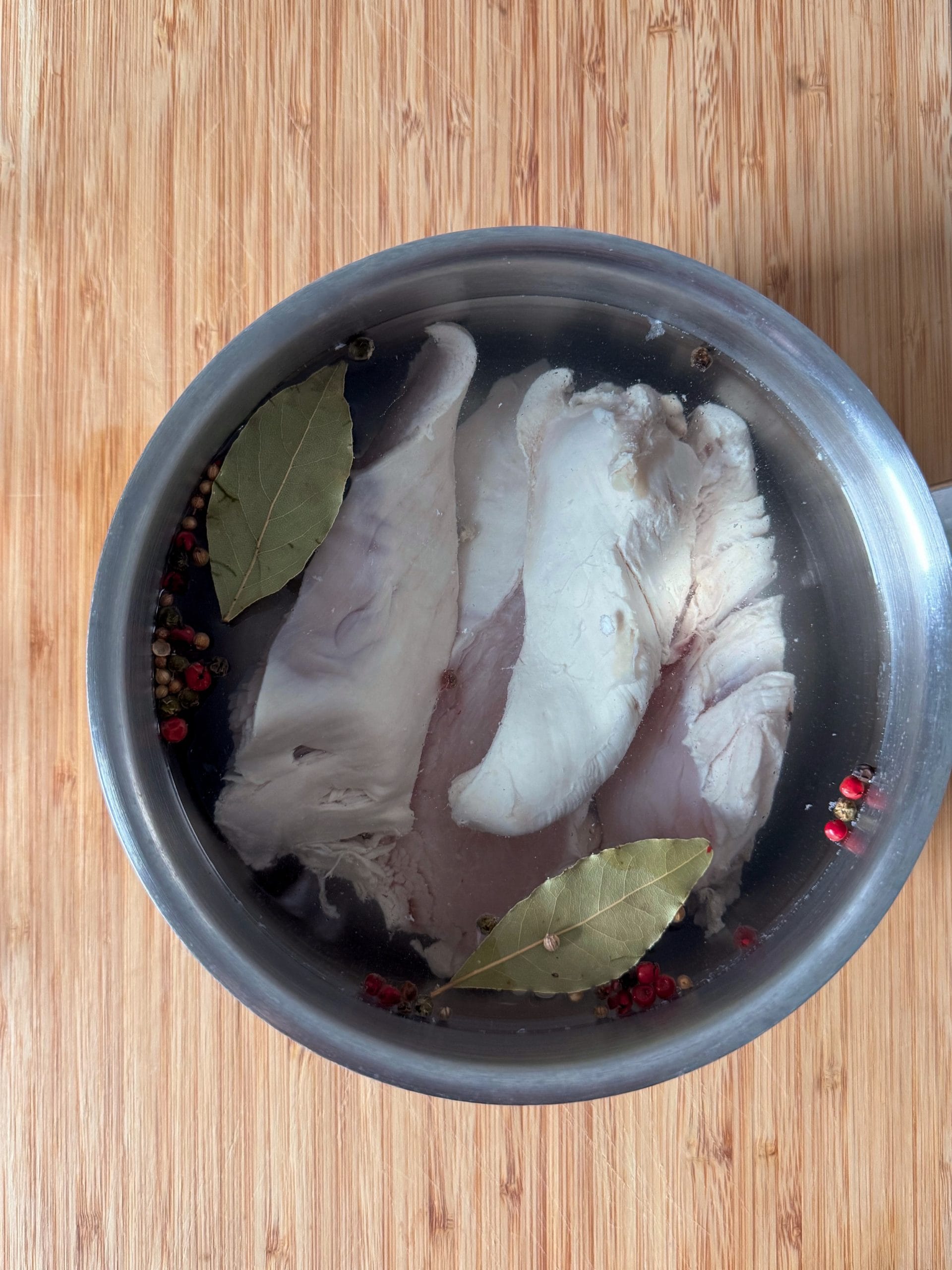 Raw chicken breast in a pot of water with bay leaves and colorful peppercorns, ready to boil