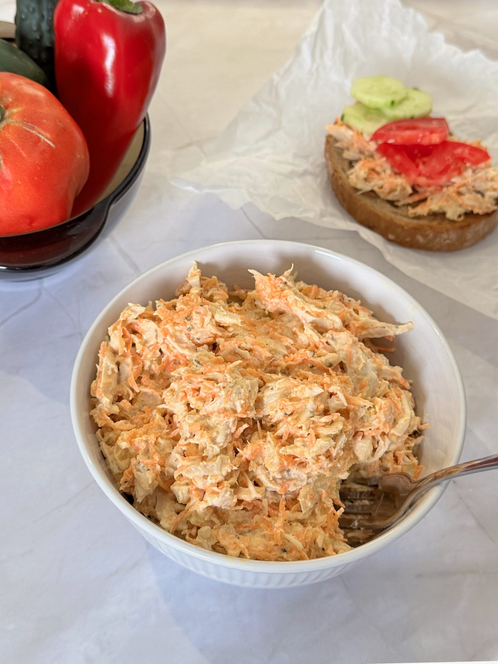 Chicken and carrot salad with Greek yogurt in a white bowl, with tomatoes, bell pepper and cucumbers in the background