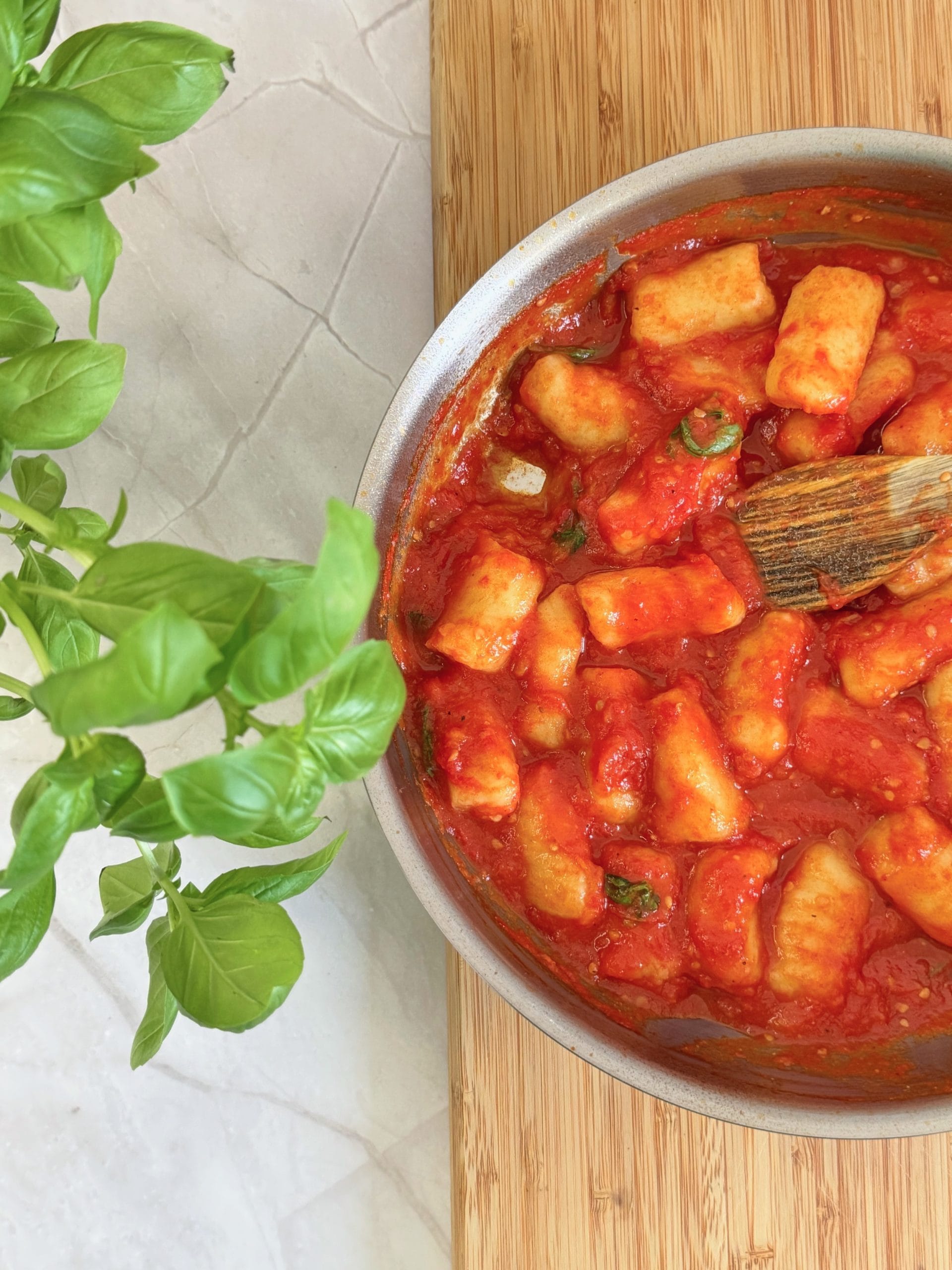 Gnocchi amestecați cu o lingură de lemn într-o tigaie cu sos de roșii și busuioc, lângă un ghiveci cu busuioc proaspăt