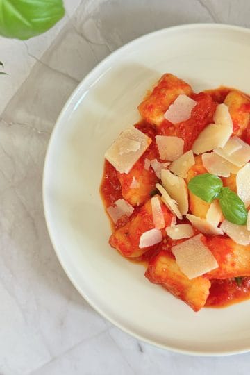 Bol cu gnocchi în sos de roșii, presărat cu fulgi de parmezan și frunze de busuioc proaspăt, pe un blat de marmură
