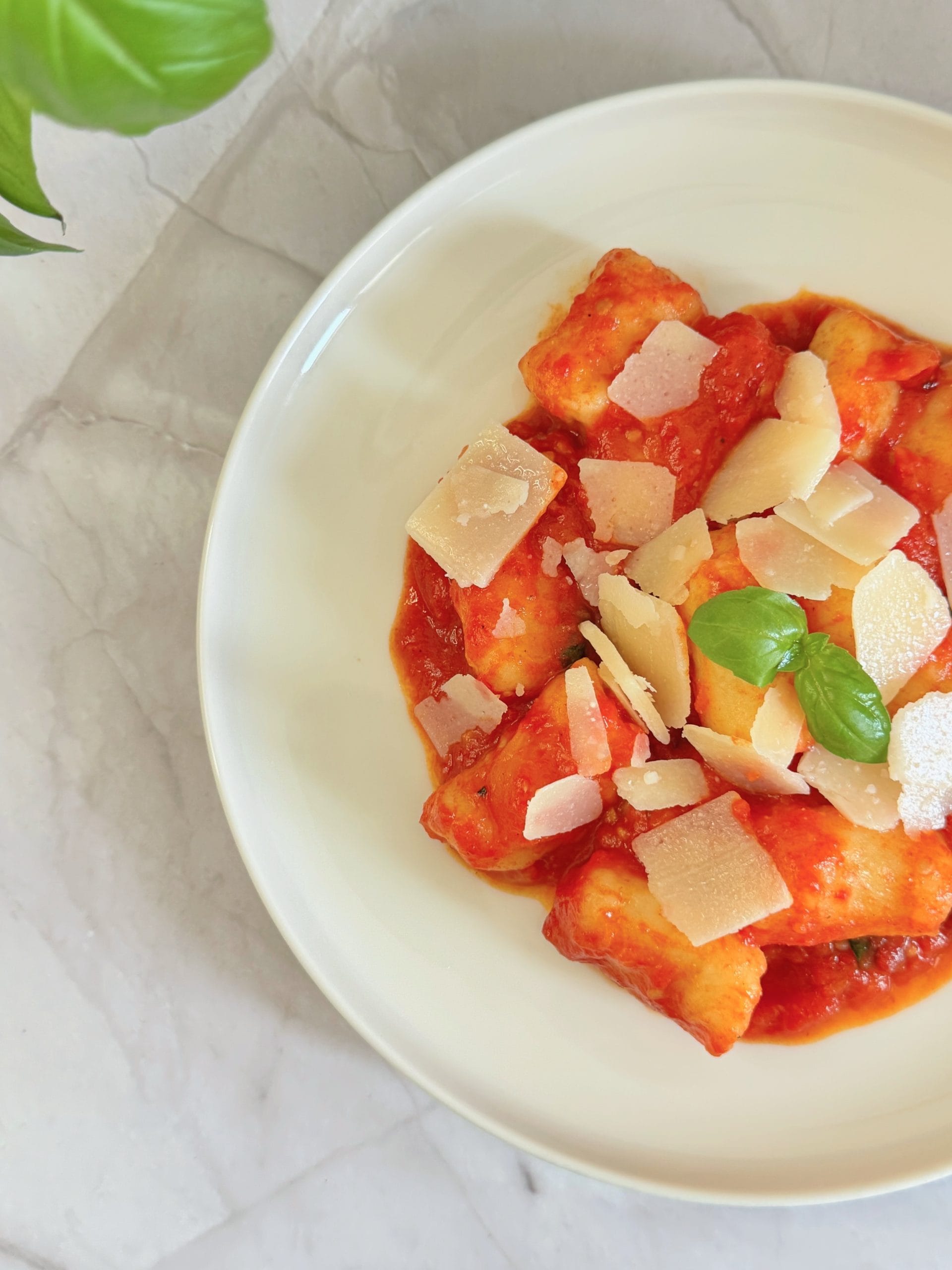Bol cu gnocchi în sos de roșii, presărat cu fulgi de parmezan și frunze de busuioc proaspăt, pe un blat de marmură