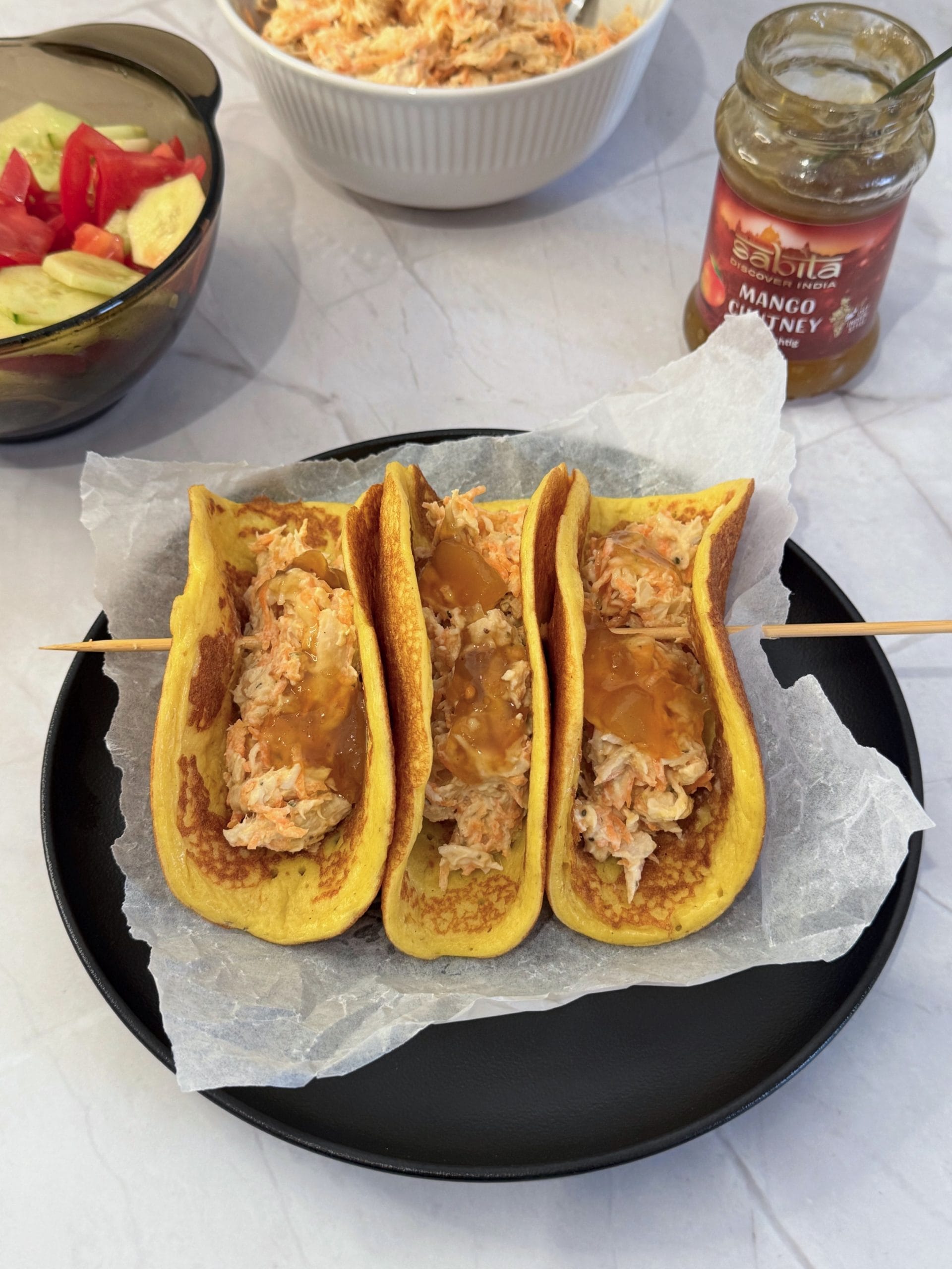 Trei lipii proteice &icirc;ndoite ca tacos, umplute cu salată de pui cu morcov și chutney de mango, pe o farfurie neagră cu h&acirc;rtie de copt, cu un borcan de chutney și salata de castraveți cu roșii &icirc;n fundal