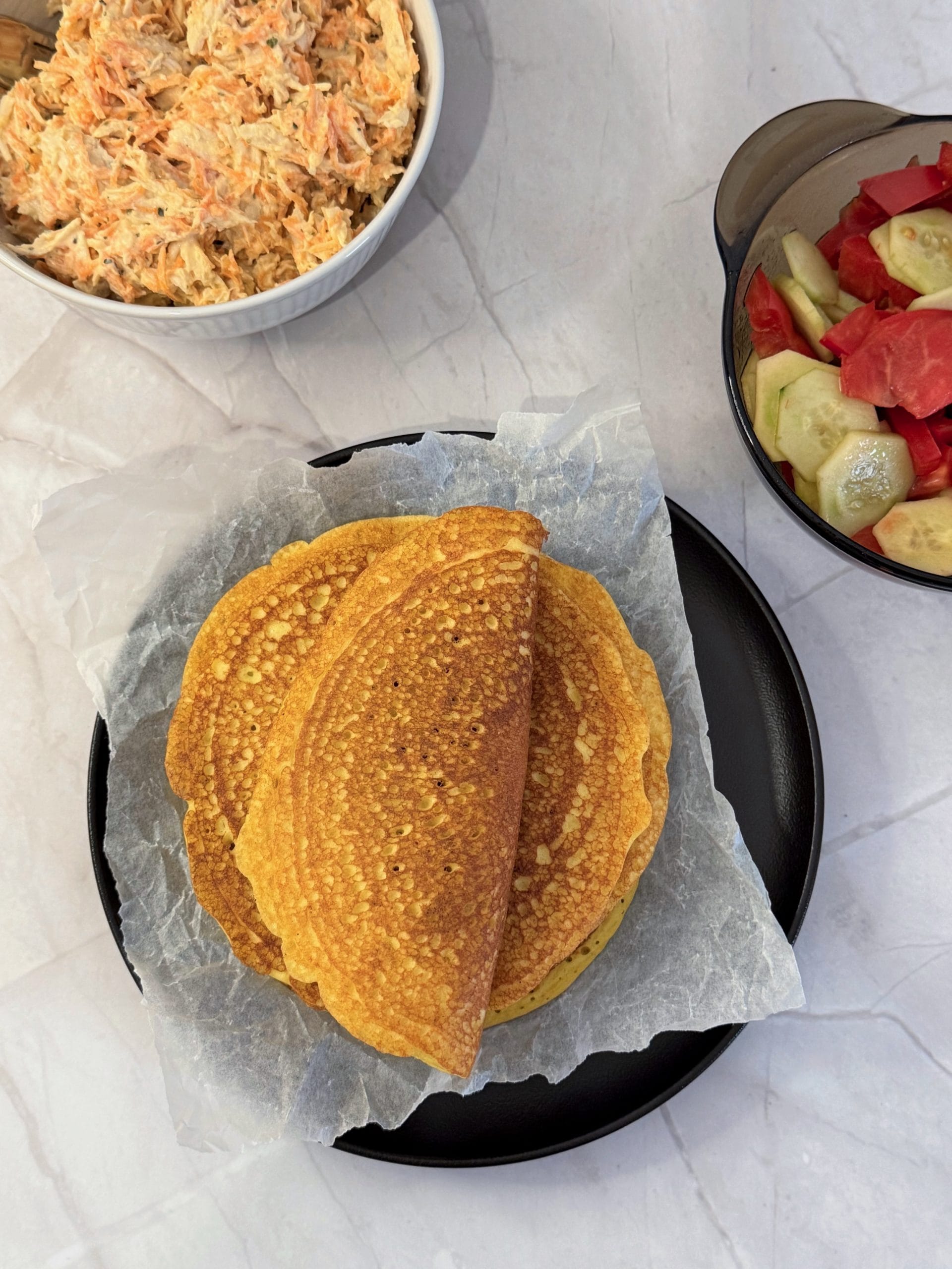 Vedere de sus cu un teanc de lipii proteice aurii pe h&acirc;rtie de copt, pe o farfurie neagră, una &icirc;ndoită &icirc;n jumătate, l&acirc;ngă un bol cu salată de pui cu morcov și un bol cu salată de castraveți și roșii