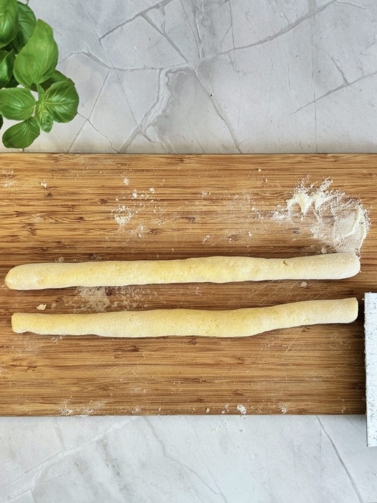 Două suluri de aluat de cartofi pentru gnocchi, rulate pe un blat de lemn presărat cu făină, lângă frunze de busuioc și un liniar