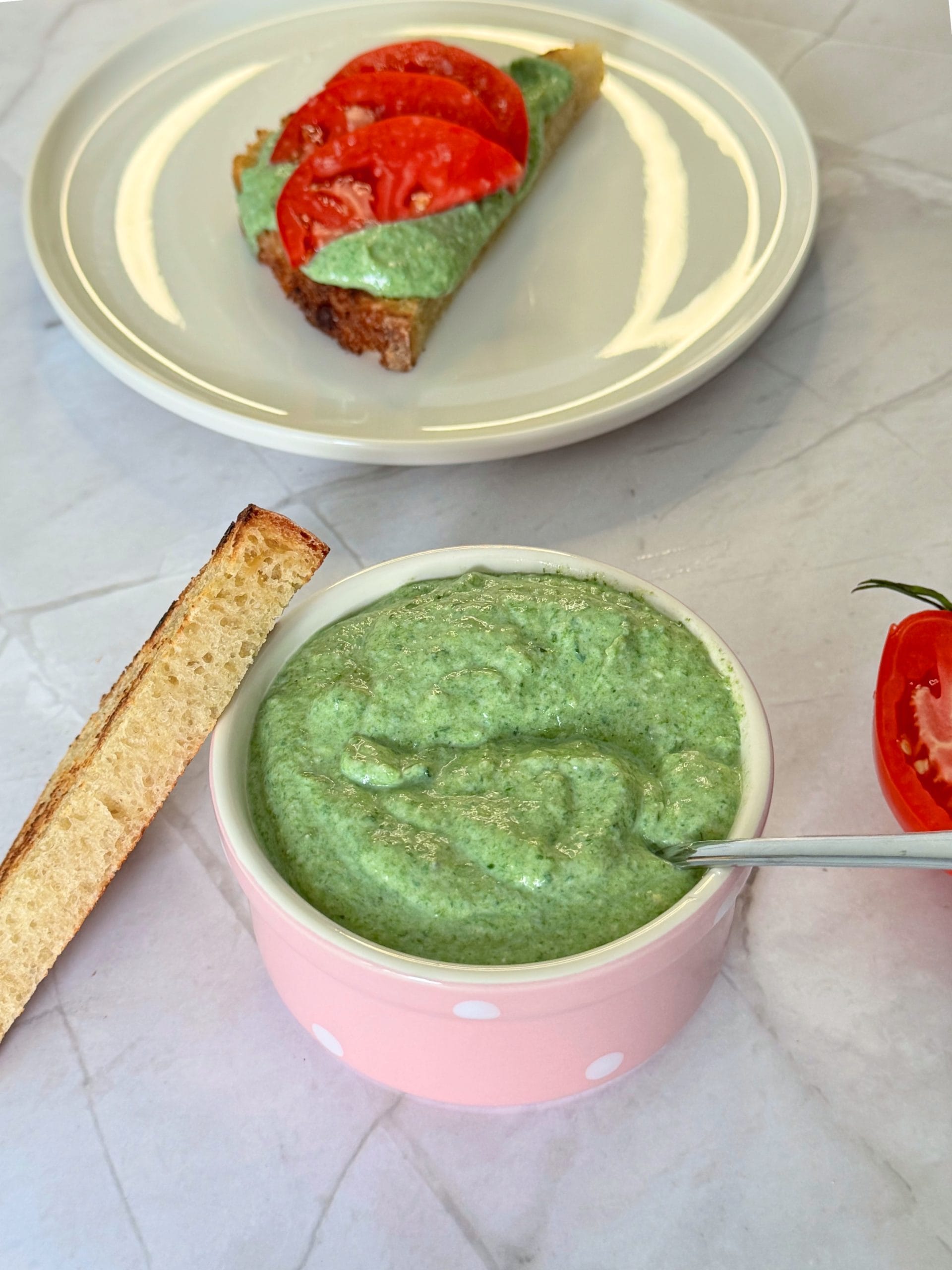 Spinach ricotta cream in a pink bowl, next to a slice of toast topped with the cream and sliced tomatoes