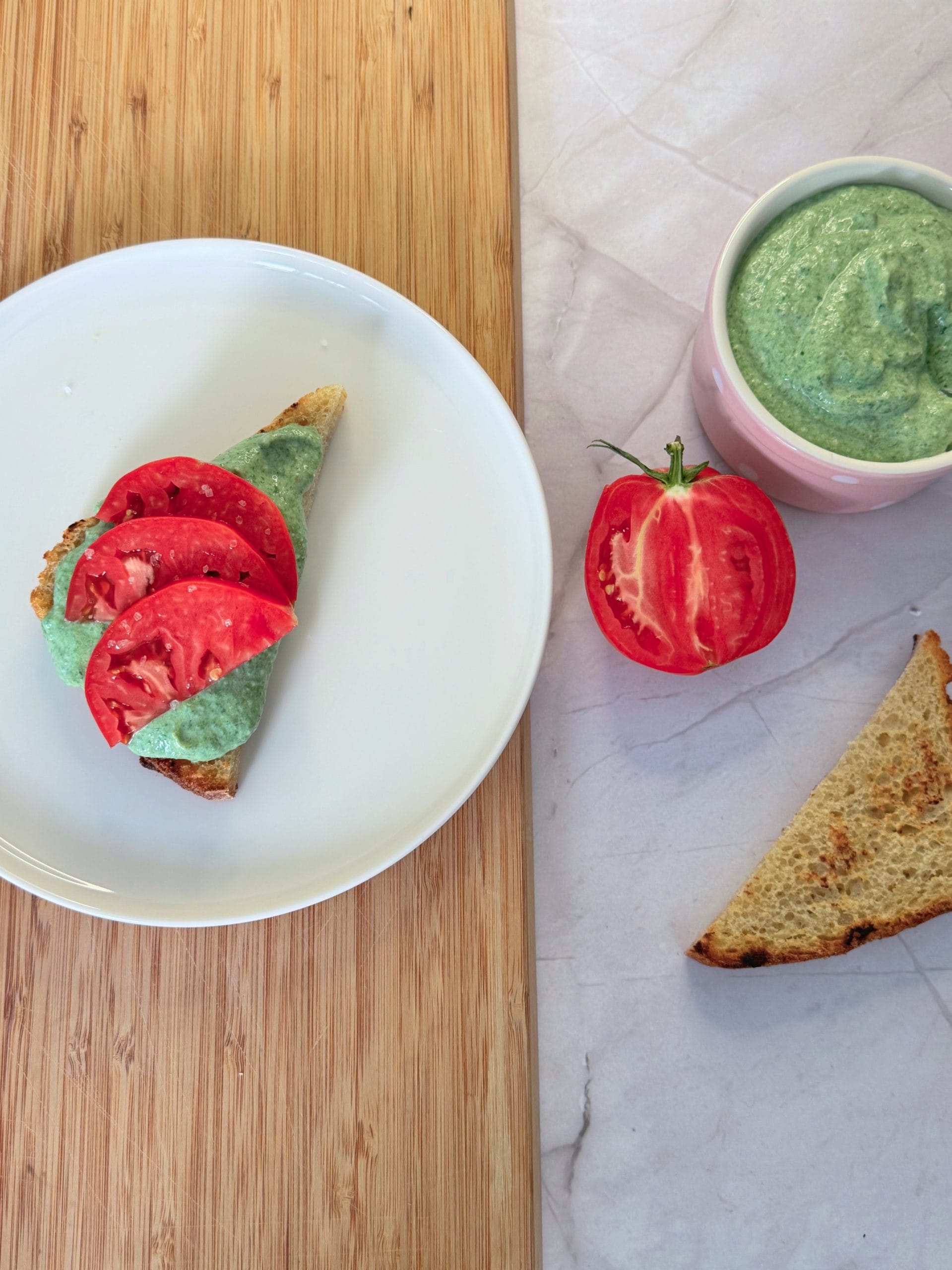 Slice of toasted bread topped with green spinach ricotta cream and tomato slices, served on a white plate