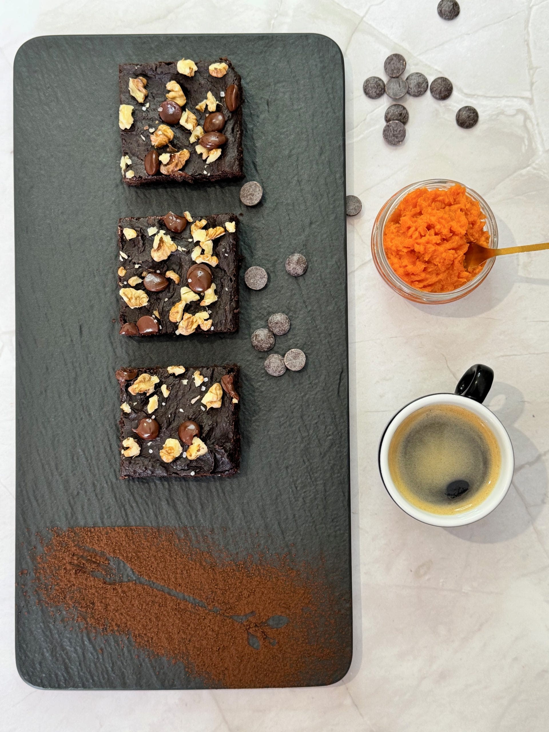 Trei felii de brownie cu cartof dulce pe platou de ardezie, cu nuci, ciocolată și sare, fotografiate de sus lângă o cafea espresso și un bol cu piure de cartof dulce