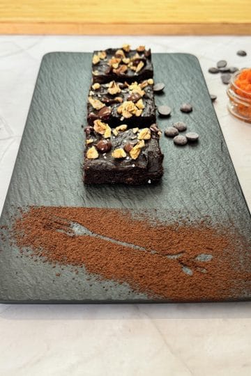 Sliced sweet potato brownie topped with walnuts, chocolate and sea salt, arranged on a slate platter dusted with cocoa, next to mashed sweet potato and coffee
